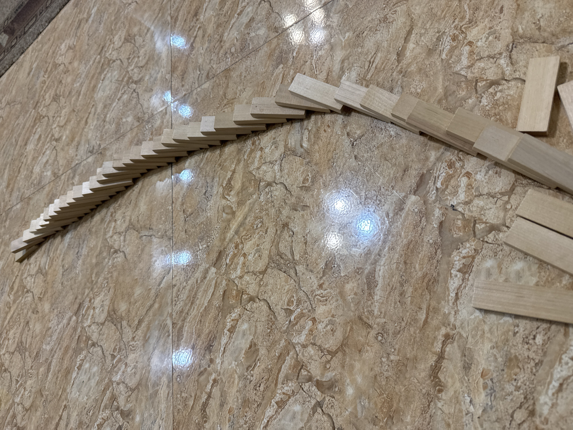 The image shows a series of wooden dominoes set up in a curved line on a tiled floor. The tiles are brown and have a marbled pattern. The dominoes start in the upper left corner and curve toward the lower right corner. Some dominoes are standing and some are not. There is no indication of the location in Yerevan, Armenia, or any people present in the image.
