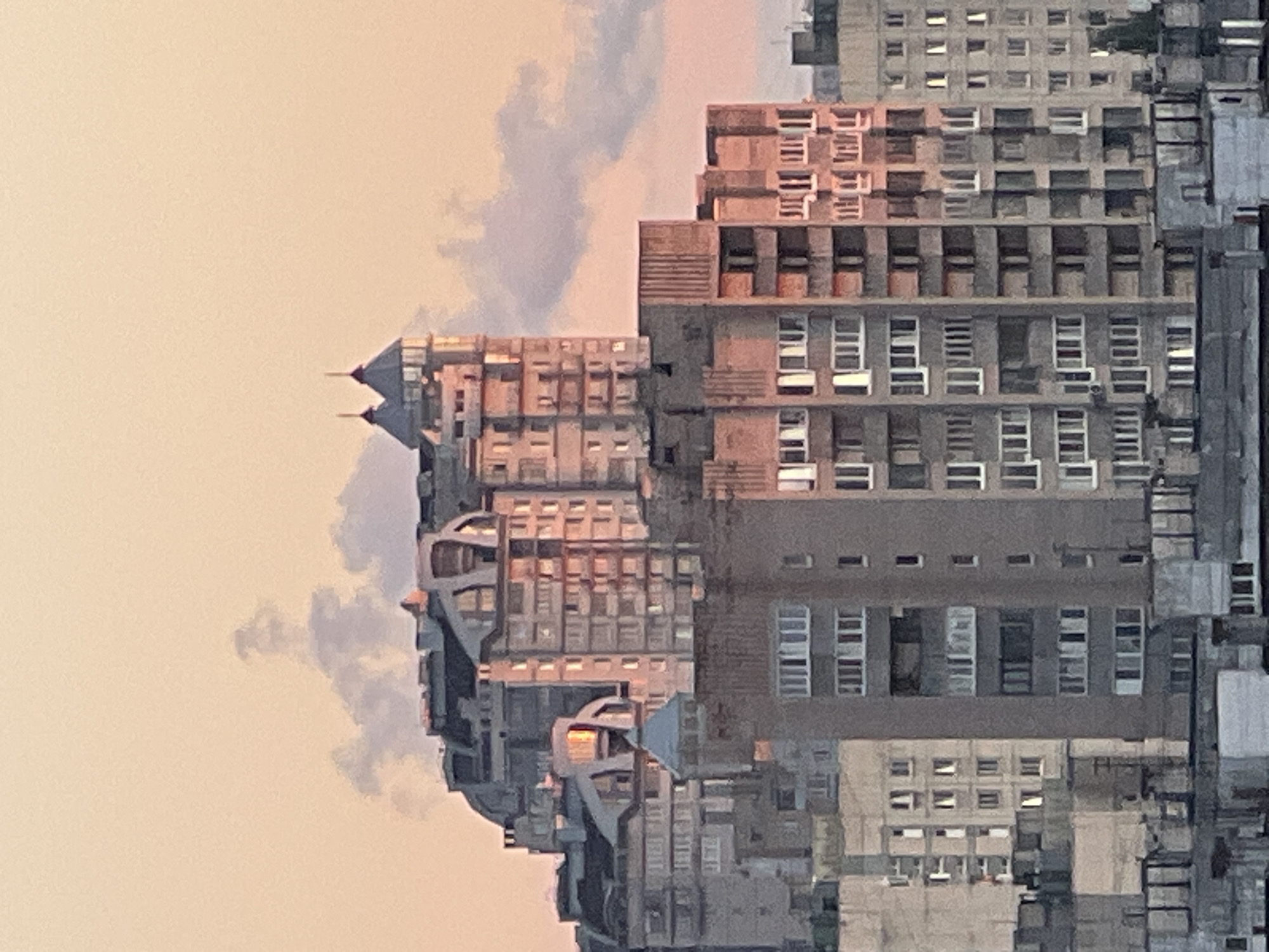 The image captures an urban landscape dominated by a dense cluster of apartment buildings at dawn or dusk. The sky is a soft, peachy-orange hue, with wispy clouds tinged with pink. The buildings, varying in height and architectural style, are constructed from concrete and display a uniform, greyish facade, with numerous windows. Some of the buildings feature modern, rounded designs with ornate tops, while others are stark, geometric structures. Sunlight catches some of the upper levels, casting a warm glow on the concrete. The overall impression is one of a vast residential area.