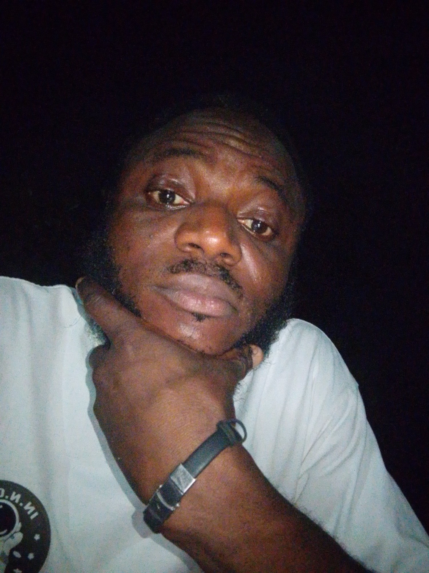 A dimly lit, close-up portrait features an adult man of color, captured at night or in a very low-light environment. He is shown from the chest up, looking directly towards the camera with a neutral to slightly serious expression. The man has dark skin, a full beard, and a mustache. His left hand is raised, with his index finger and thumb resting thoughtfully against his chin and jawline. He is wearing a white t-shirt, and a dark-colored wristband with a silver clasp is visible on his left wrist. On the left side of his t-shirt, a circular logo or patch is partially visible, featuring text that appears to include "O.N.NI" and a graphic with a potential astronaut or space motif. The background is almost entirely dark, concentrating the focus on the man's face, which is illuminated, causing his eyes to appear somewhat reflective. The image exhibits some graininess, characteristic of photography taken in challenging low-light conditions. The photograph was taken in Lokoja, Nigeria.