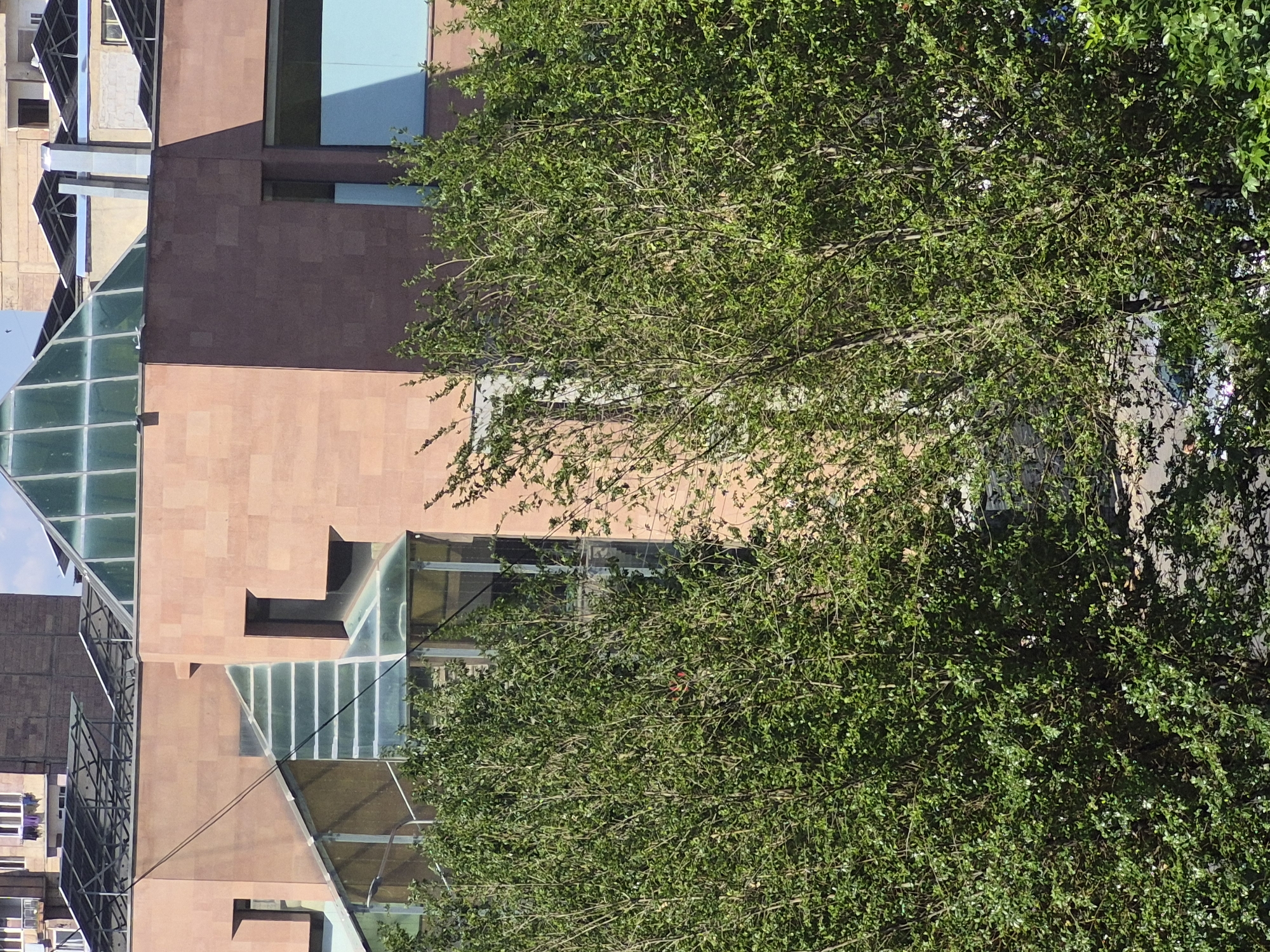 The image displays a modern architectural scene in an urban environment, likely in Yerevan, Armenia, with a significant portion of a contemporary building obscured by a dense, leafy green tree.
The building features a multi-story design characterized by a combination of reddish-brown and lighter tan stone or paneling. Its most striking elements are the extensive angular glass sections, which form sloped roofs and walls, reflecting the bright blue sky and surrounding structures. These glass elements often have a visible grid-like framework. Rectangular windows are integrated into the solid facades, and distinct light blue rectangular panels or windows are visible on the upper parts of the structure, adding a splash of color. Another, older-style building with a darker roof and more traditional windows can be seen in the background or adjacent to the main building.
In the foreground and covering much of the right side, a large, lush tree with numerous small green leaves and visible branches creates a natural screen, with sunlight filtering through its foliage.
The scene is captured during the daytime under bright, clear conditions, evidenced by the strong shadows cast by the building and the tree. No people or specific activities are depicted. There is no visible text within the image.