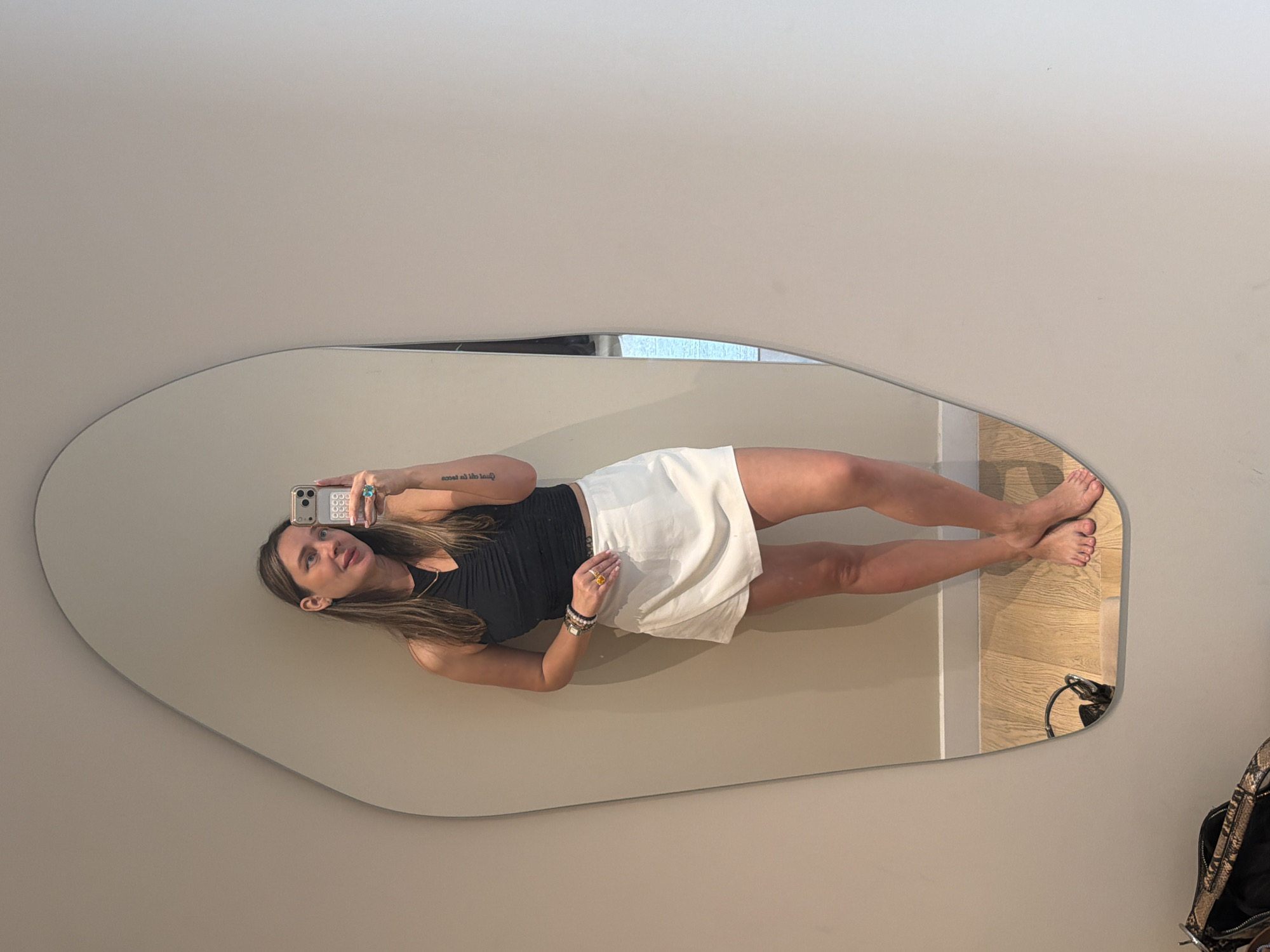A woman stands in front of a large, irregularly shaped mirror, taking a selfie. She is wearing a black ruched halter top and a white A-line mini skirt. She has long, light brown hair and is wearing bracelets and rings. Her left arm is tattooed. She is barefoot, and her feet are visible in the mirror reflection on a wooden floor. A black handbag with a snakeskin pattern is on the floor in the lower left corner of the image. The background is a plain, light-colored wall. The scene appears to be indoors, possibly in a dressing room or apartment. There is no indication of the location being in Moscow, Russia.