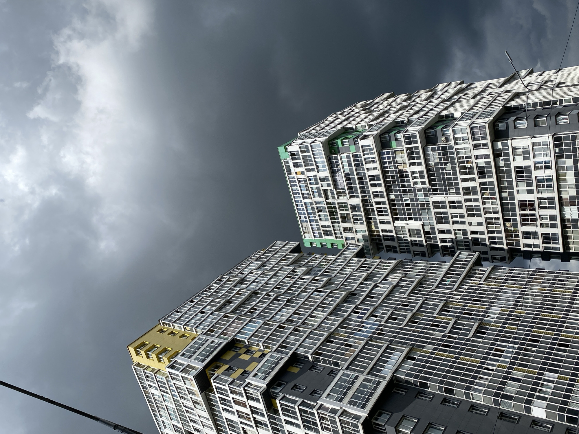 Two modern, high-rise residential buildings stand against a dramatic, overcast sky. The building on the left is predominantly white and grey with yellow accents on its upper floors and balconies. The building on the right features white and grey exteriors with vibrant green sections on its upper levels and balconies. Both buildings have numerous windows, creating a grid-like pattern across their facades. In the upper left quadrant of the image, a portion of a dark, slender pole, possibly a streetlamp or utility pole, extends upwards. The sky is filled with dark, heavy clouds, suggesting an impending storm. The scene is a view of urban architecture in Kudrovo, Russia.