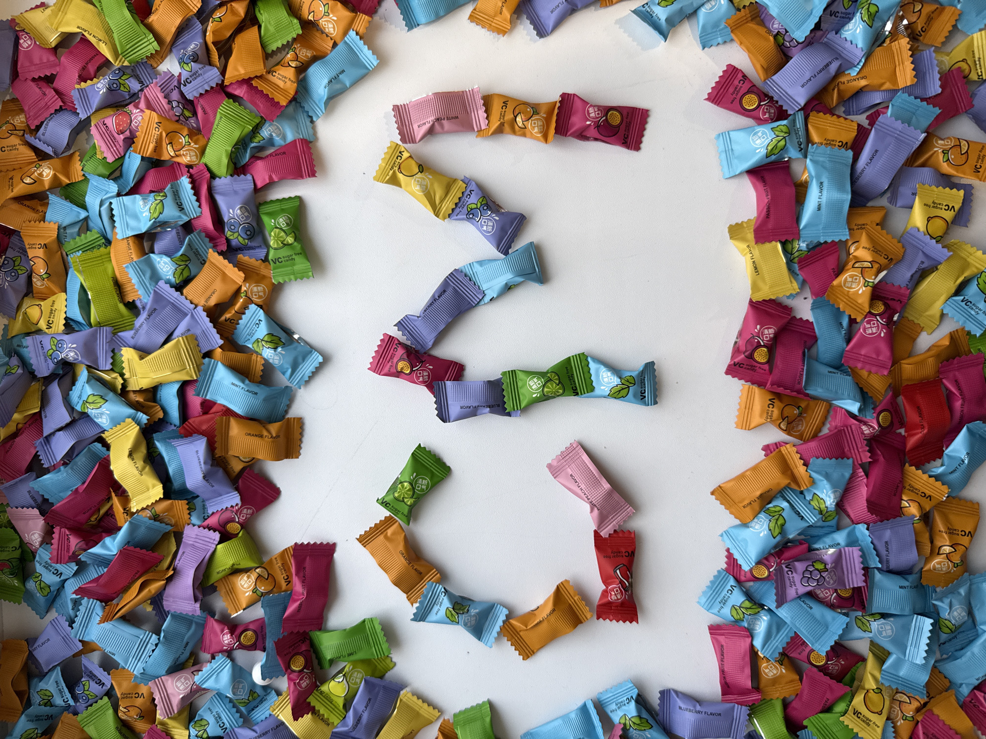A top-down shot reveals a white surface scattered with numerous individually wrapped candies. The candies, likely hard candies based on their angular shape, come in a variety of colors, including yellow, orange, pink, purple, blue, red, and green. Each wrapper features an image of a fruit and text. The candy wrappers are piled densely at the edges, but are arranged in the center to outline a shape resembling the number "3".
