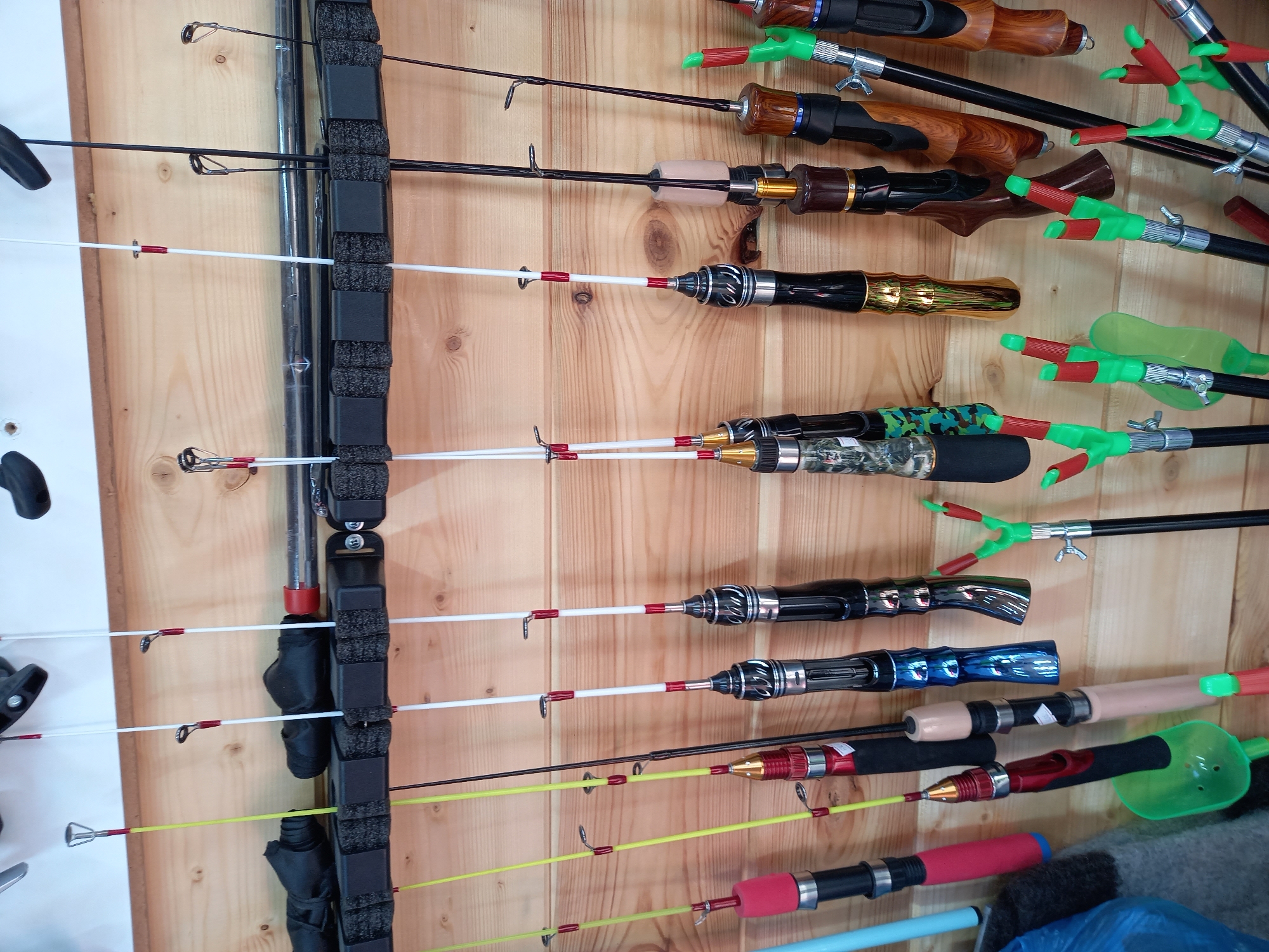 The image shows a collection of fishing rods mounted on a wooden wall. On the left side, rods are held in a black rack with foam inserts. The rods have various colors and handle styles, with some having plastic rod holders attached. The background is a light-colored wooden surface.