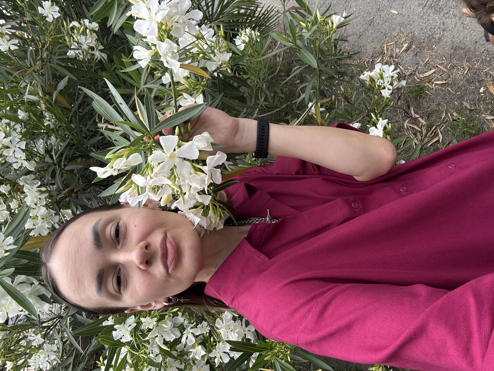 A young woman with dark hair and light skin is standing in front of a bush with white flowers. She is wearing a bright pink collared shirt and a silver necklace with a cross pendant. She has a silver hoop earring with a cross charm in her left ear and a black smartwatch on her right wrist. Her right hand is holding a stem of the flowering bush. The background is a dense green bush with many white flowers. The ground is visible in the lower right corner, showing a dirt path with fallen leaves. The scene appears to be an outdoor environment, possibly a park or a garden, in the city of Sochi, Russia.