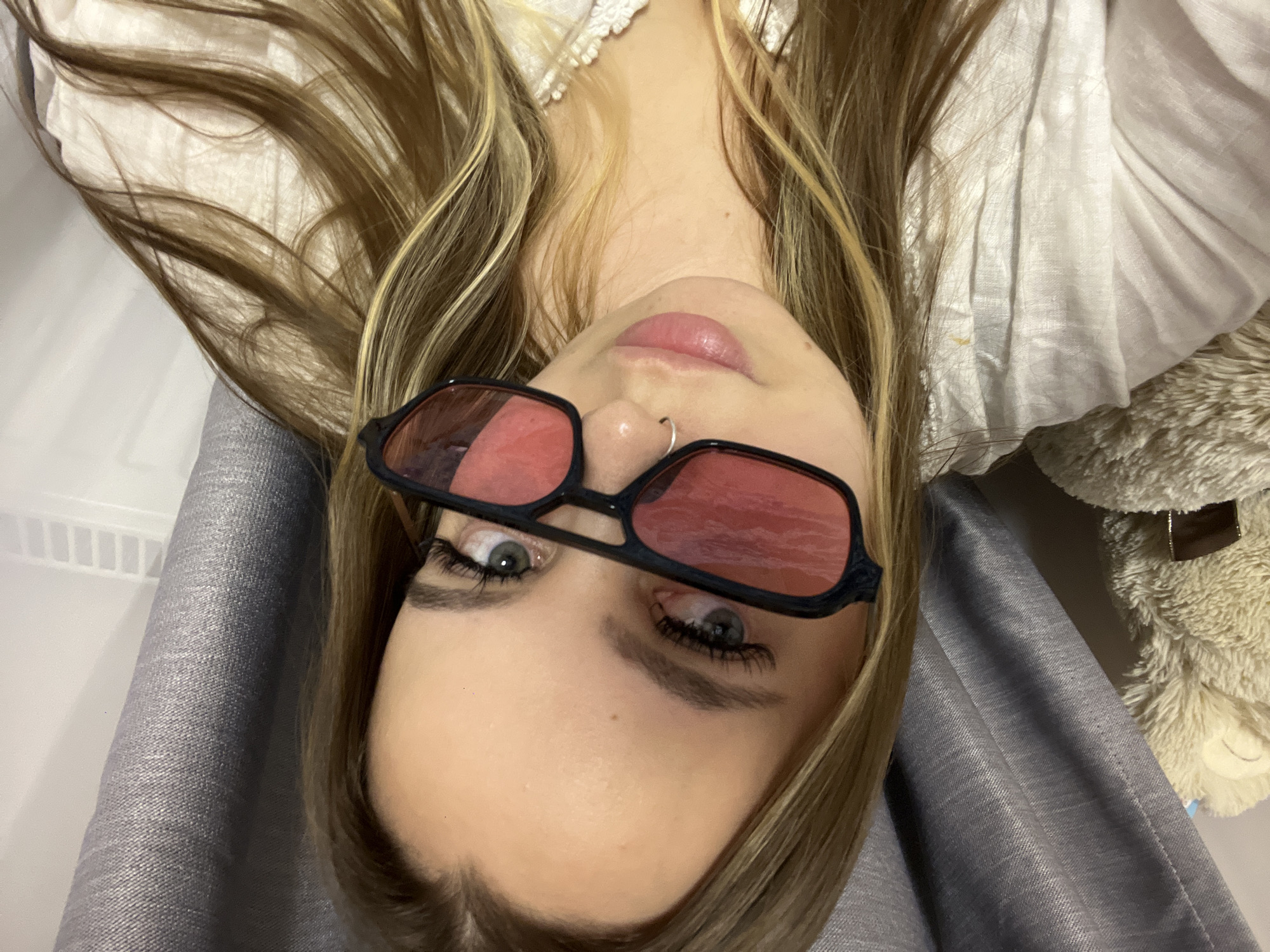 This is a close-up, top-down shot of a young woman, likely taken as a selfie or from an overhead perspective, with her head appearing inverted relative to the frame. The scene is set indoors, in Krasnodar, Russia.
The woman is the central subject, with long, wavy hair featuring a mix of blonde highlights and brown tones, fanned out around her head. She wears a pair of black-framed, rectangular sunglasses with red-tinted lenses, through which her light-colored eyes (appearing green or blue) are visible. Her eyelashes are prominent, and she has a small silver hoop nose ring. Her lips are full and pink.
She is wearing a white top or dress with a delicate, possibly ruffled or textured neckline. A grey, woven fabric, which could be part of her clothing or a blanket, is visible on her left side and shoulder. To her right, a portion of a fluffy, light-brown teddy bear or stuffed animal can be seen. The background appears to be a light-colored surface, possibly bedding or a wall, with a white grid-like structure (potentially a vent or shelf) in the top left corner. The lighting is soft and even, suggesting an indoor environment.
The woman is looking directly at the camera, posing for the photograph. There is no visible text in the image.