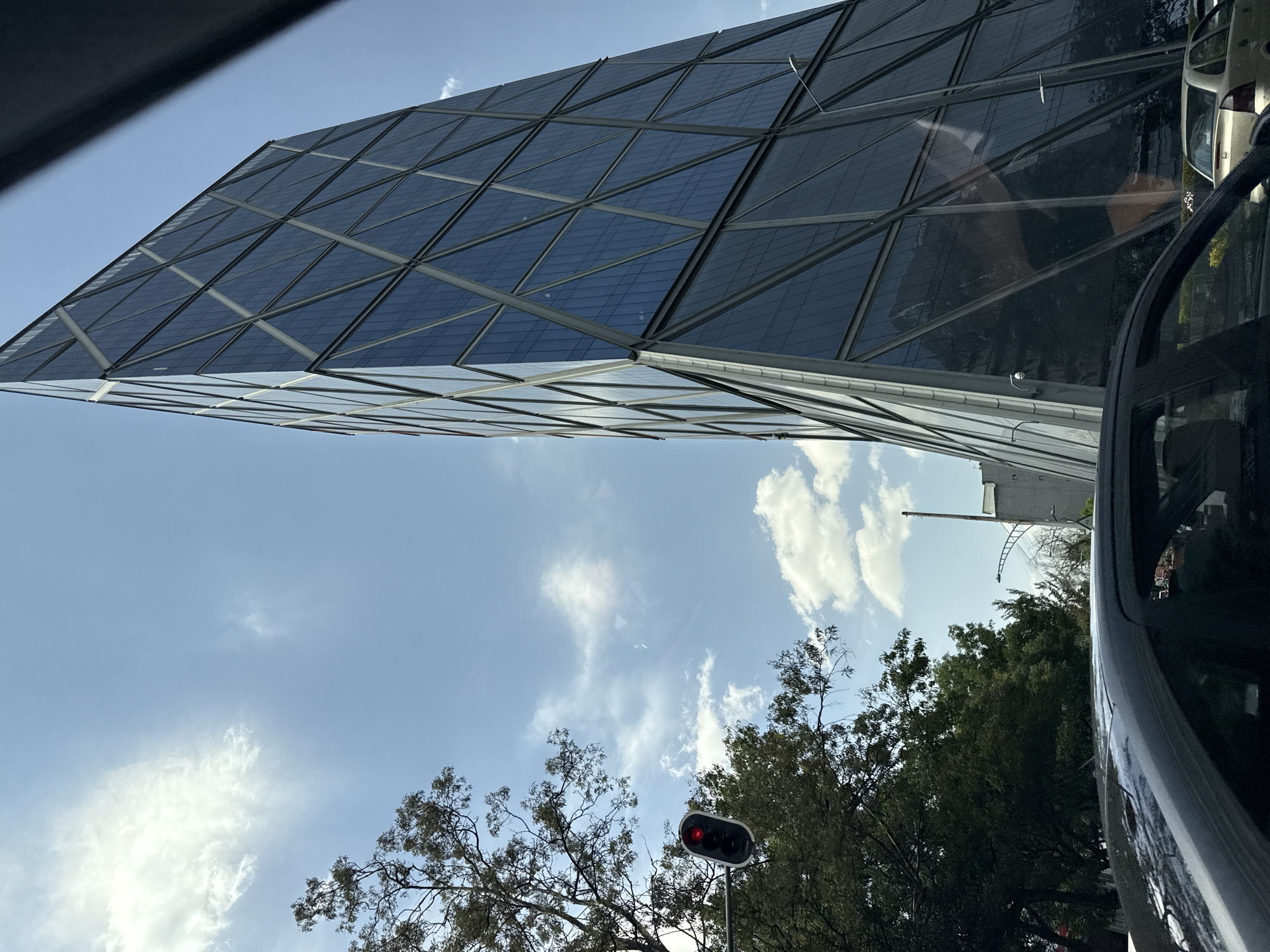 The image shows a view from inside a vehicle in Mexico City, Mexico. A distinctive glass building with a triangular grid pattern dominates the upper portion of the frame. The sky, with scattered clouds, provides a backdrop to the building. In the lower portion of the frame, treetops and a traffic light displaying a red signal are visible. Part of the vehicle’s interior is also visible on the right side of the frame.