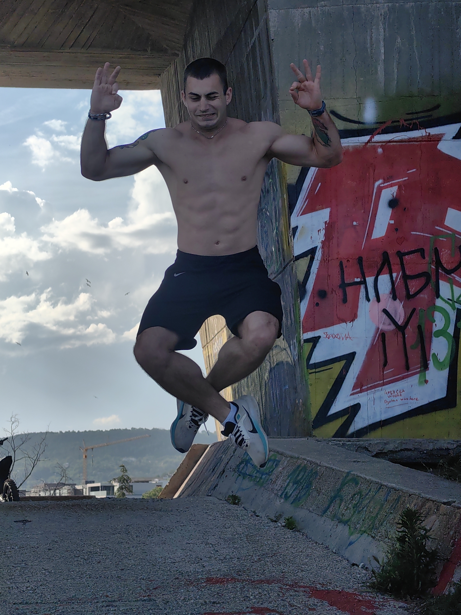 A young man is captured mid-air, jumping over a concrete structure covered in graffiti. He is shirtless, wearing black shorts and athletic shoes. His arms are raised, with his fingers forming a peace sign on each hand. The background features a cloudy sky and a large concrete wall adorned with vibrant graffiti, some of which appears to be in Cyrillic script. In the distance, a construction crane is visible, suggesting an urban or developing area. The foreground consists of a textured concrete surface. This scene is located in Varna, Bulgaria, specifically near the area of the old Varna Thermal Power Plant, known for its abandoned structures and street art.