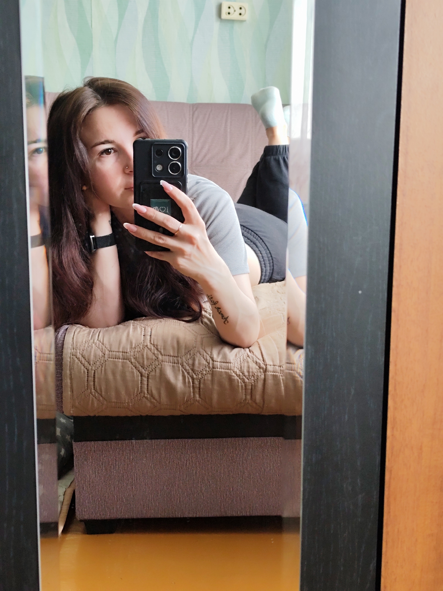 A woman is taking a selfie in a mirror. She is lying on a brown upholstered couch with a quilted texture. She is wearing a gray shirt, black shorts, a black watch, and a ring. Her hair is long and brown. She has a tattoo on her left forearm. A single leg with a white sock is sticking up. The background features a light green and white patterned wallpaper and an electrical outlet. The reflection in the mirror shows another partial view of the woman. The room has a wooden floor.
There is insufficient information to determine that the image was taken in Фоки, Russia.