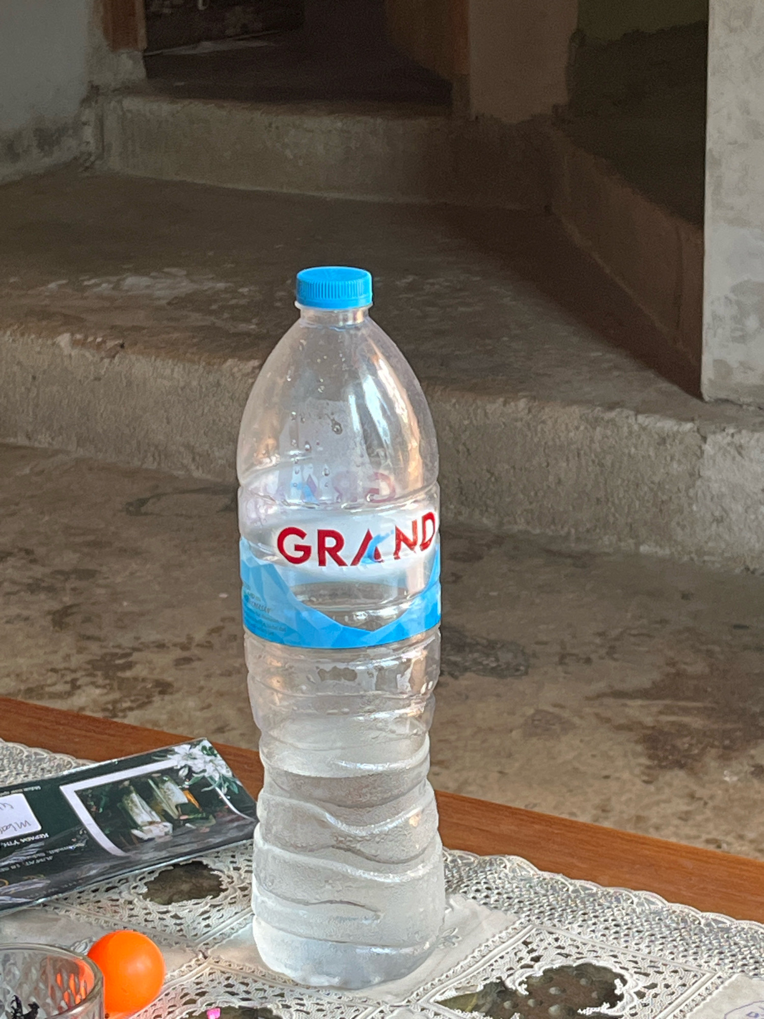 A clear plastic bottle of water with a blue cap and a label that reads "GRAND" is placed on a wooden table covered with a white lace placemat. Next to the bottle is a pamphlet or brochure featuring an image of birds. An orange ball is also visible on the placemat. In the background, a set of concrete stairs leads up to a dark opening. The overall scene appears to be outdoors, possibly on a patio or in a courtyard, with a rustic and weathered ambiance. The city of Sinar Mulyo, Indonesia, is not discernible from the image content.