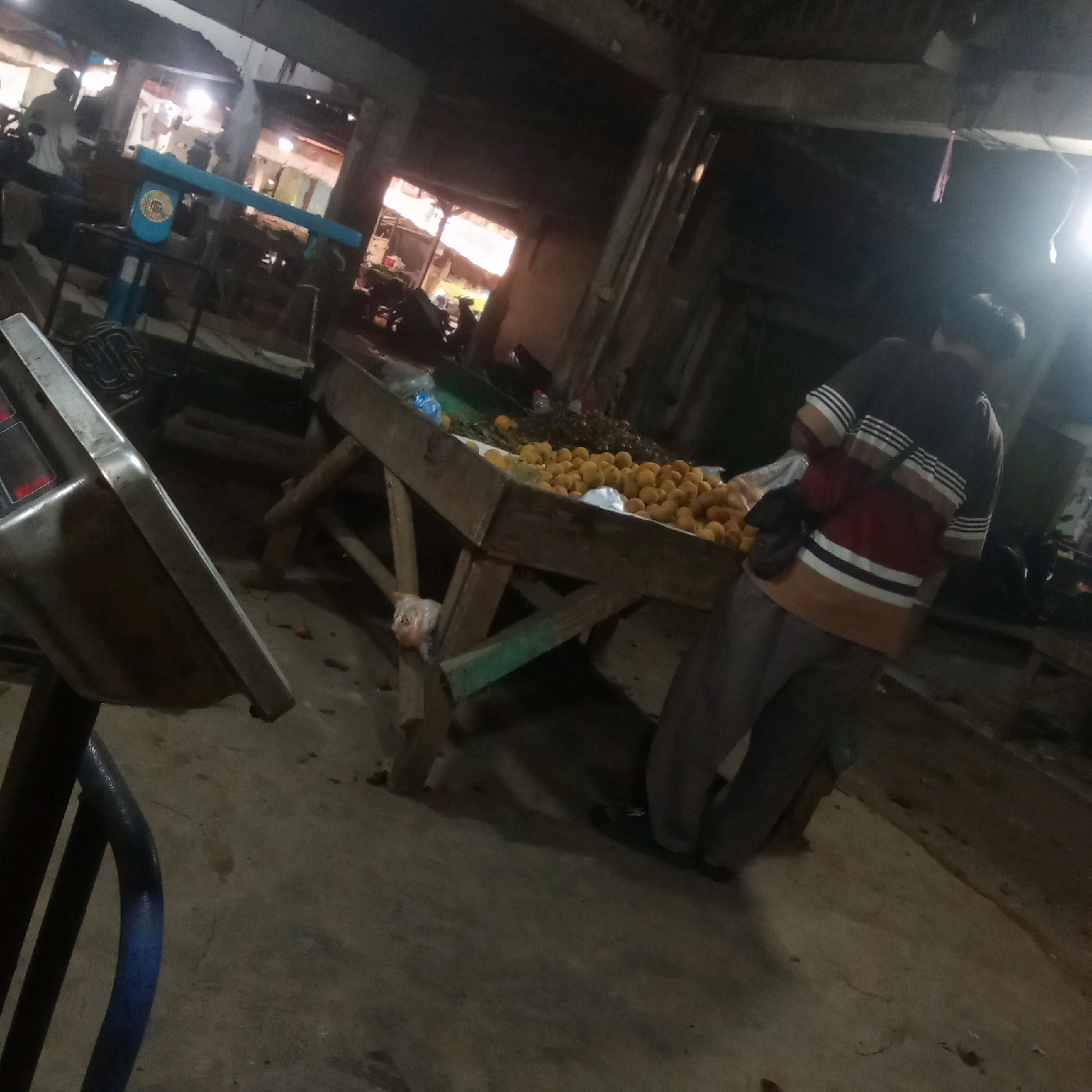 This image depicts a dimly lit indoor market scene, likely in Karawang, Indonesia, characterized by a concrete floor and overhead structures that suggest a marketplace. In the foreground, a portion of a metal weighing scale with a blue handle is visible on the left. A person, viewed from the back, is standing in the mid-ground, wearing a striped t-shirt and dark pants, with a bag slung over their shoulder. They appear to be looking towards a wooden stall laden with a large quantity of orange fruits, possibly oranges or tangerines. To the left of the fruit stall, another individual can be partially seen near a blue scale or weighing apparatus. The overall atmosphere is one of commerce, with goods displayed on stalls and people engaged in market activities, despite the low lighting conditions.