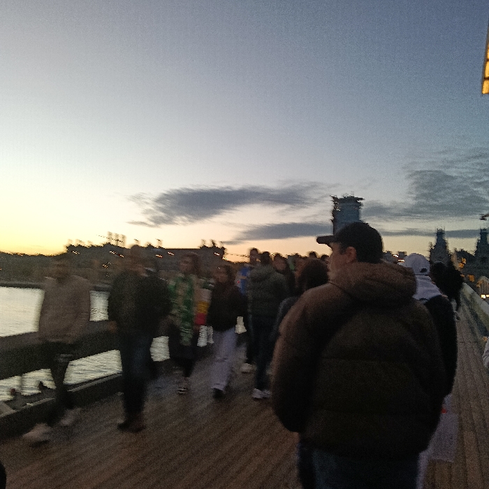 People walk on a wooden pier at dusk. The sky fades from orange to blue. Water is visible on the left. Buildings and other structures line the horizon. The crowd is diverse in age and attire, including a man in a brown puffer jacket and baseball cap dominating the foreground. The photo is somewhat blurry, suggesting movement.