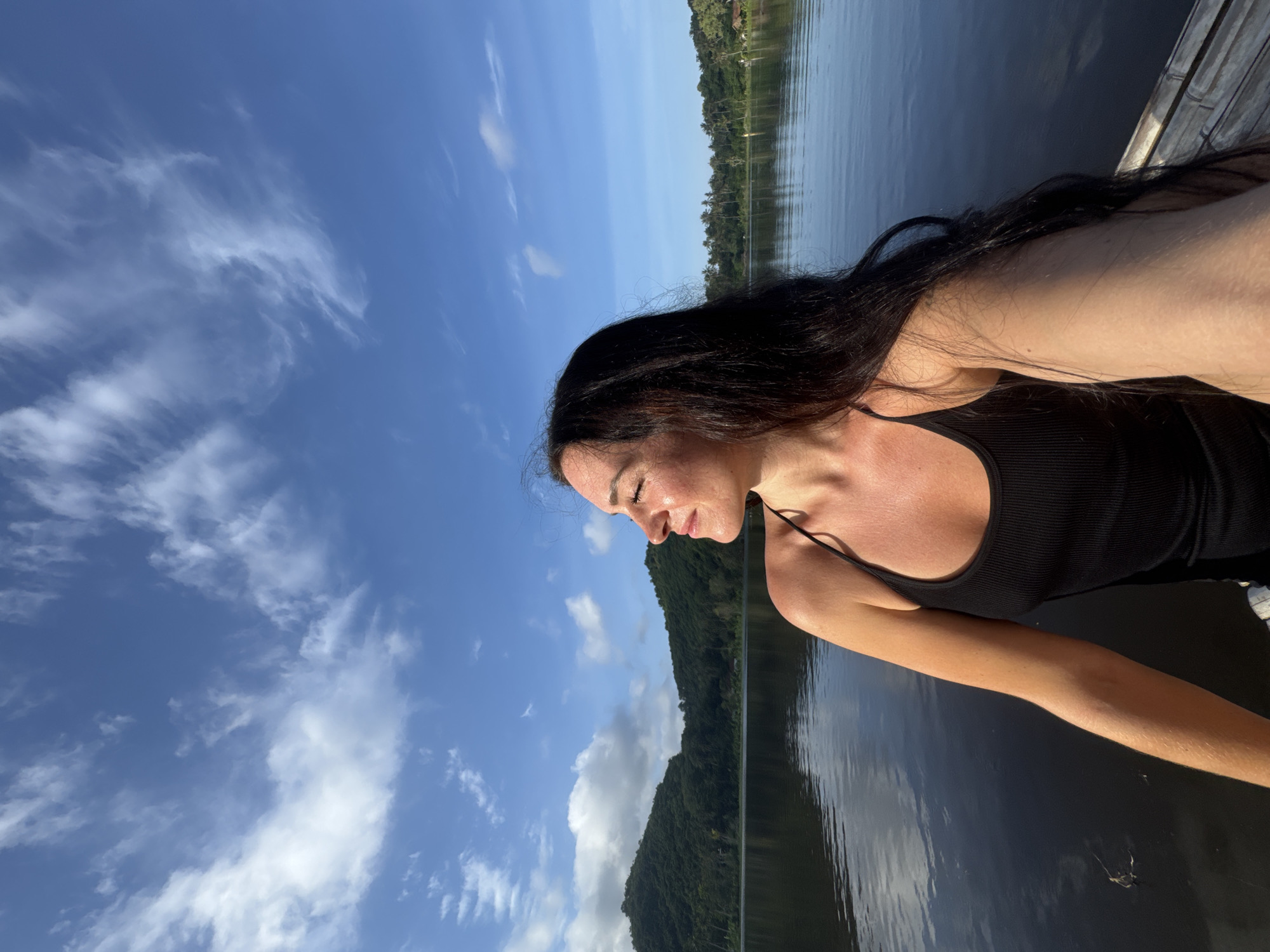 A woman with long, dark hair is pictured from the chest up, facing the right side of the frame with her eyes closed and head tilted slightly upwards. She is wearing a black tank top. She appears to be on a body of water, possibly a lake, given the calm surface and reflections of the sky and surrounding greenery. In the background, a lush green hillside covered in trees rises from the water's edge. The sky above is a vibrant blue with scattered white clouds, suggesting it is daytime and possibly morning or late afternoon due to the quality of light. The overall scene conveys a sense of peace and natural beauty.