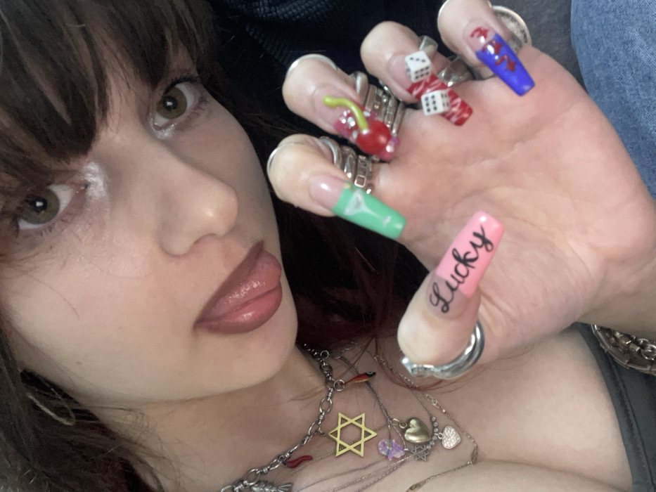 A woman with bangs and brown hair poses in a selfie. She has light skin, brown eyes, and glossy lips. Her tongue is slightly visible. The woman wears layered necklaces, one featuring a star of David. Her hand is prominently displayed with multiple rings and long, decorated nails. The nails are colorful, including one with a cherry, dice, and the word "Lucky." The background includes glimpses of a black garment and blue denim.