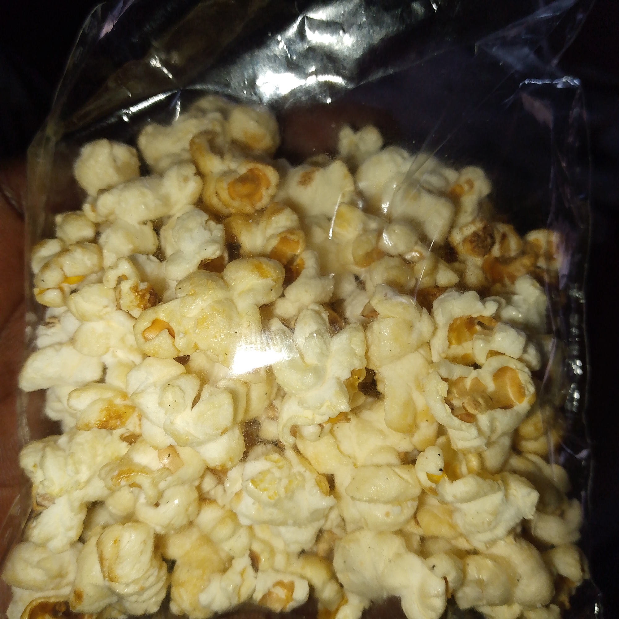 A close-up, high-angle shot features a clear plastic bag filled with popcorn. The bag is mostly full of white and off-white popped kernels, with several pieces displaying golden-brown and slightly darker, caramelized sections, indicating a potential sweet or buttery flavoring. Some kernels appear to be partially popped or retain a darker, unpopped core. Small dark specks, possibly seasoning, are scattered among the popcorn.
The clear plastic packaging is crinkled, particularly at the top, and shows distinct reflections from an unseen light source, creating bright, elongated highlights across its surface. A dark, out-of-focus background surrounds the bag, emphasizing the brightly lit popcorn within. There is a slight hint of a hand holding the bag on the left side, blurred and indistinct. No activity is depicted beyond the static presentation of the bagged snack. The overall lighting suggests low ambient light conditions, with a direct light source illuminating the popcorn. There is no visible text or branding on the packaging.