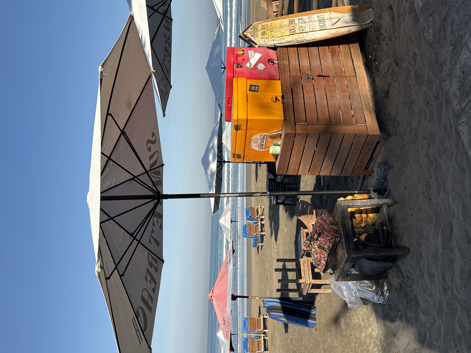 This image depicts a beach scene in Kuta, Indonesia. The beach is sandy and sunny with a clear blue sky overhead. Several beach umbrellas are visible, some with discernible advertisements. Beach chairs line the shore near the water. A weathered wooden structure sits in the foreground, topped with two ice chests, one red and one yellow, and a sign with handwritten text. Next to the wooden structure is an open box containing coconuts. Some other items lie on the sand, including a plastic bag and beach gear. The overall atmosphere is relaxed and typical of a tourist beach location.