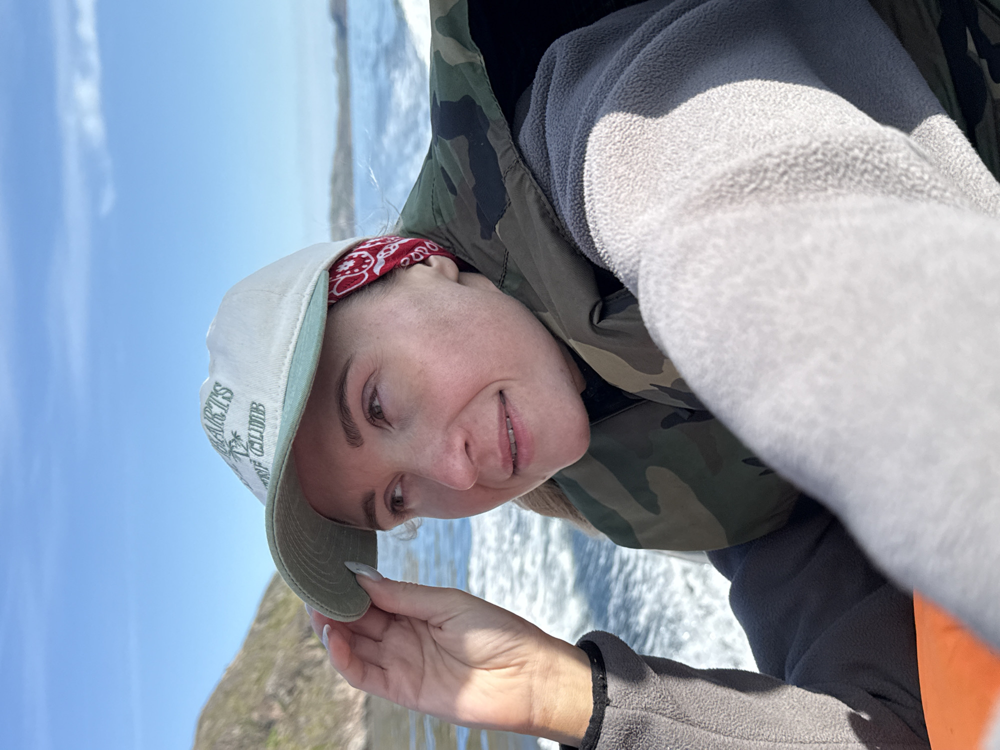 In the city of Teriberka, Russia, a woman is photographed from the chest up while on a boat. She is wearing a white baseball cap with a green brim, a red bandana, a camouflage life vest, and a grey fleece jacket. Her left hand is holding onto the brim of her cap, and she is looking to her right. The background shows a body of water with waves and a distant shoreline under a clear blue sky.