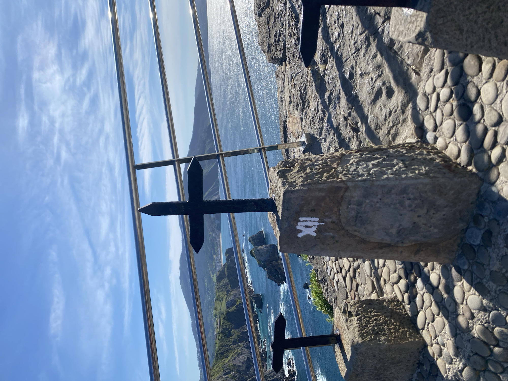 The image depicts an outdoor scene on a sunny day, likely in the vicinity of Bermeo, Spain, given the coastal landscape. In the foreground, three cross-shaped markers are visible, each mounted on a stone pedestal. The central cross is black, with a larger base and pointed top, and has the Roman numeral "XII" painted in white on its pedestal. To the left, another black cross is partially visible, also with a pointed top and mounted on a square stone base. A third cross is suggested to the right, also on a stone base, but mostly out of frame.
The ground is paved with smooth, rounded pebbles. Behind the crosses, a metal railing with multiple horizontal bars runs across the scene, separating the foreground from the background. The background features a rugged, green coastline with rocky outcrops jutting into a deep blue sea. The water is choppy, with white waves breaking against the rocks. Further in the distance, rolling hills covered in vegetation are visible, with some structures and a town nestled along the coast. The sky is a vibrant blue with scattered wispy white clouds. The sunlight casts strong shadows on the paved ground.