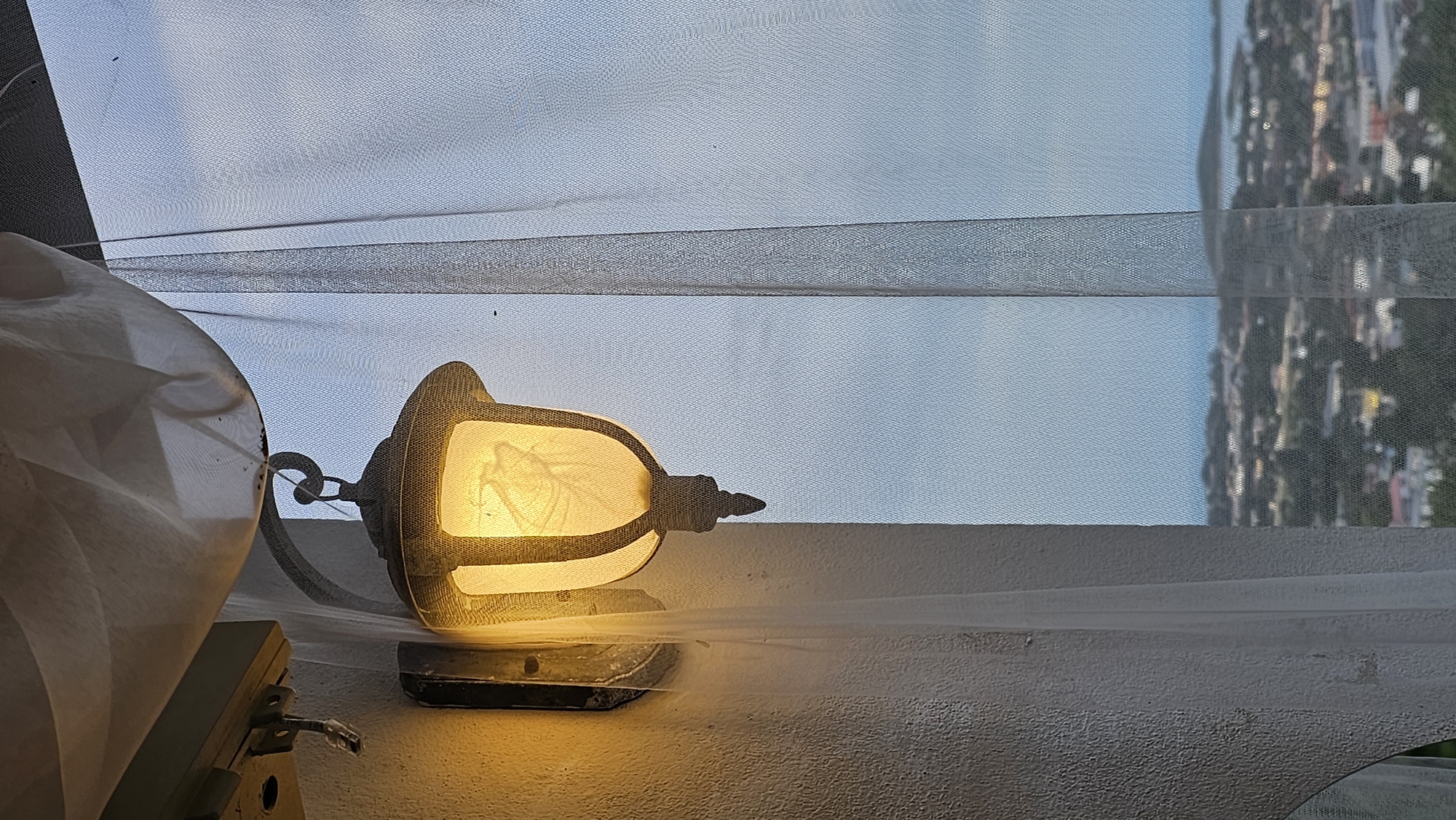 The image depicts an outdoor scene at dusk, possibly from a balcony or window in Baan Chaweng, Thailand. The main subject is an illuminated outdoor lantern, casting a warm yellow glow. Inside the lantern's glass, a delicate, translucent object resembling a leaf or feather is visible, illuminated by the light. The lantern is mounted on a textured, light-colored wall.
To the left, a large, cream-colored, draped object, possibly fabric or a lampshade, occupies a significant portion of the foreground. A hook and part of a mounting bracket are also visible beneath it, suggesting this object is also attached to the wall or a structure.
The background features a light blue sky with subtle cloud formations, indicative of either early morning or late evening. A fine mesh or screen covers the entire scene, creating a hazy effect and a subtle texture over the sky and the distant view. Through this mesh, a blurry reflection of a cityscape or densely built area with trees is visible on the right side, hinting at the location being urban or semi-urban.
The overall mood is quiet and serene, enhanced by the soft lighting and the twilight sky. No people are visible in the frame. There is no visible text. The gentle illumination of the lantern and the soft light in the sky suggest a peaceful transition from day to night.