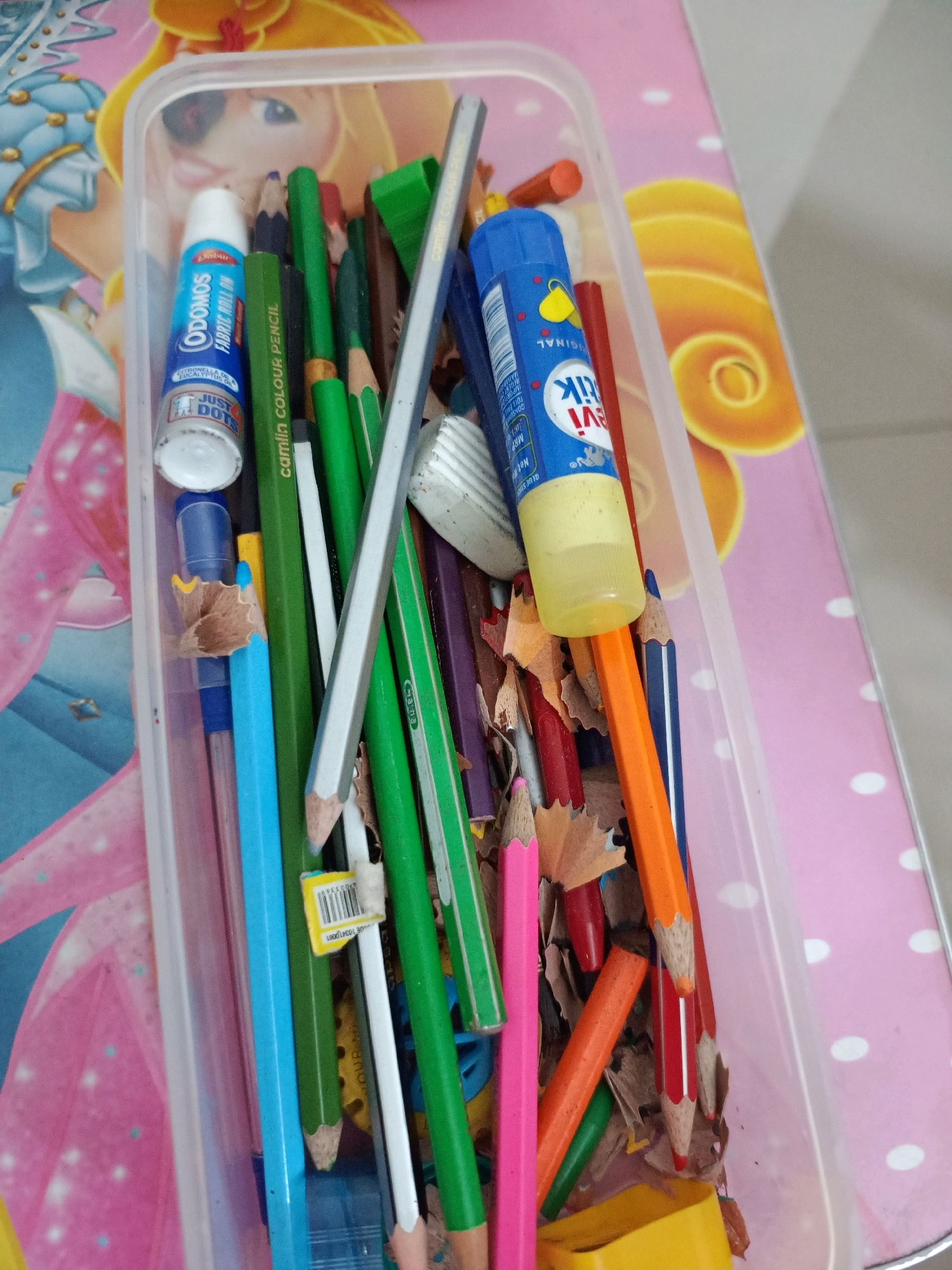 A transparent plastic container is filled with various art supplies, including colored pencils, a glue stick, and a roll-on bottle. The container is placed on a pink surface decorated with white polka dots and a pattern of yellow swirls. The background of the container is a blue and pink surface with images of cartoon characters. The scene appears to be in a domestic setting, possibly a child's art area, in Doddabele, India. The lighting suggests an indoor environment with diffused natural light.