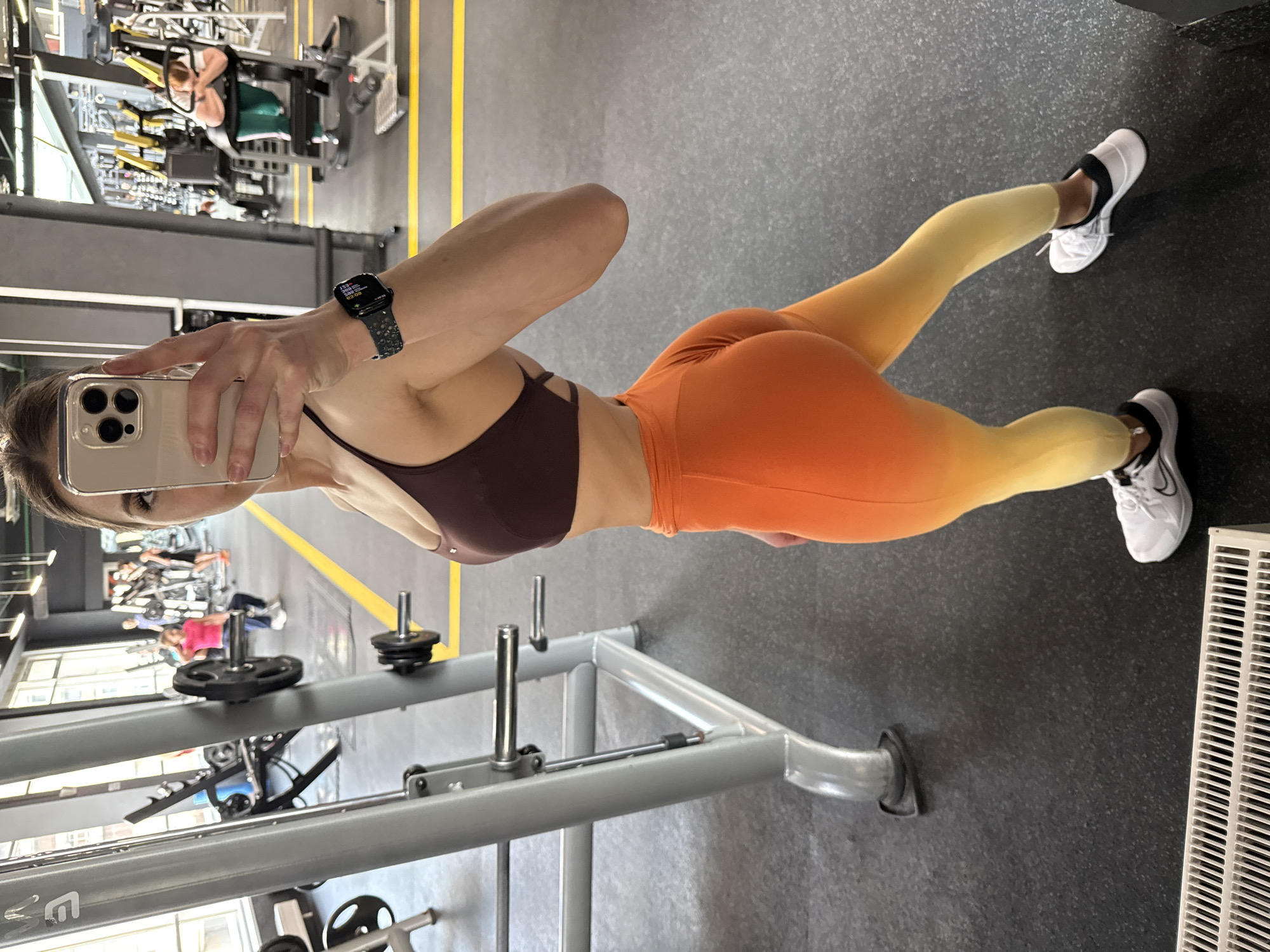 A young woman is taking a selfie in what appears to be a gym. She is wearing a brown sports bra and orange-to-yellow gradient leggings, along with white athletic shoes. She holds a smartphone in her left hand, its screen visible, and wears a smartwatch on her left wrist.
The background shows various gym equipment, including weight machines and free weights, with other people working out. The floor is dark gray, and there are yellow lines on the floor.
Given the appearance of the gym and equipment, it is difficult to specifically pinpoint the location as Saint Petersburg, Russia without further clues.