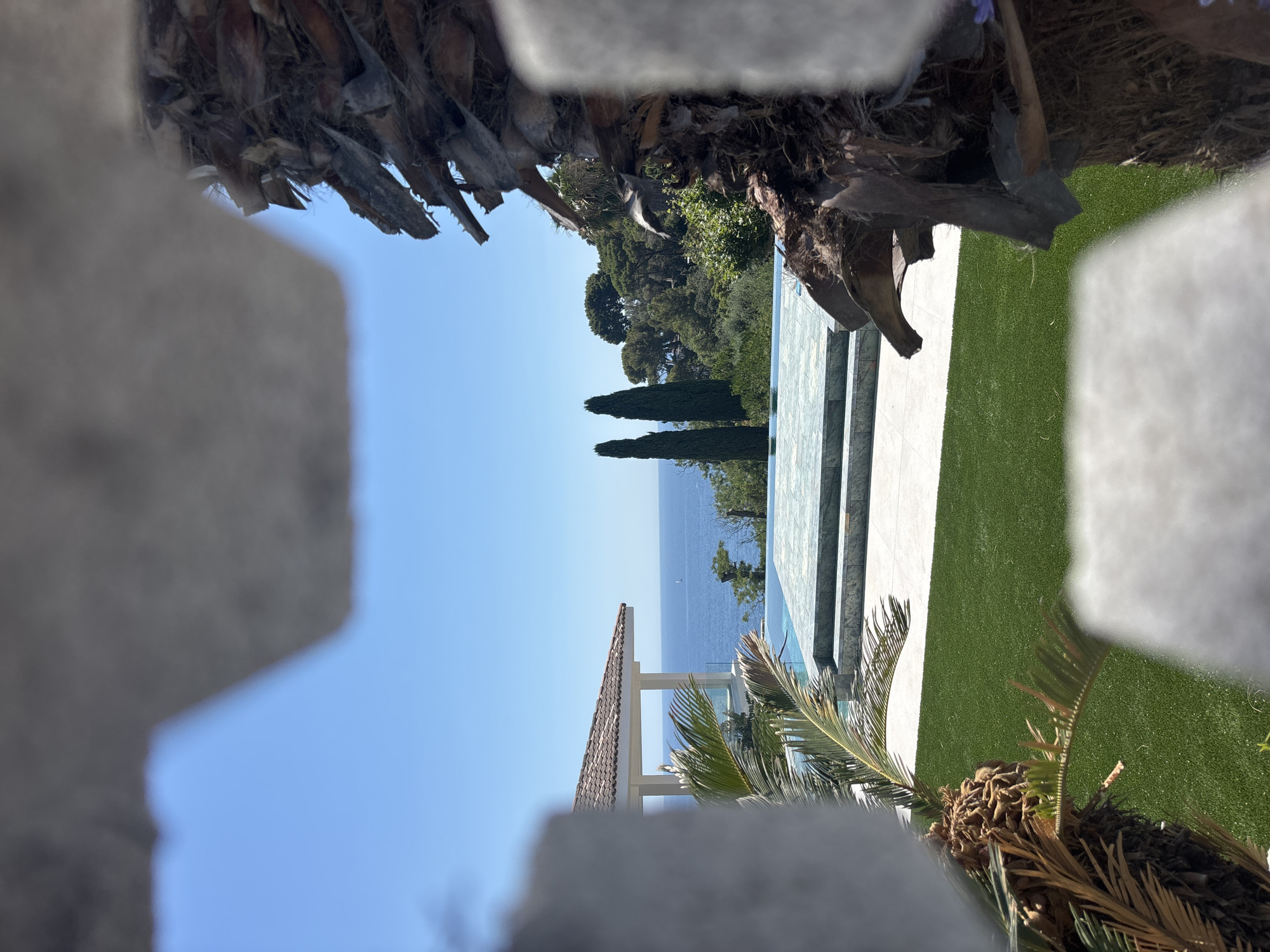 A perspective shot through the foliage of a palm tree and a light-colored rough-textured object reveals a luxurious outdoor scene on a sunny day. In the background, the deep blue Mediterranean Sea stretches to the horizon, meeting a clear, light blue sky. A small white sailboat is visible on the water.
In the midground, an infinity pool with turquoise water is framed by a tiled edge and a white tiled patio. Beyond the pool, lush green trees cover a hillside, with two tall, slender cypress trees standing prominently. A white gazebo with a terracotta tiled roof is situated to the left of the pool, offering shade and a view of the sea. Artificial green grass covers the foreground lawn.
The overall scene suggests a high-end property, likely a private residence or resort, in a Mediterranean coastal location. This view is consistent with properties found in the affluent area of Blanes, Spain, known for its beautiful coastal landscapes and villas overlooking the sea.