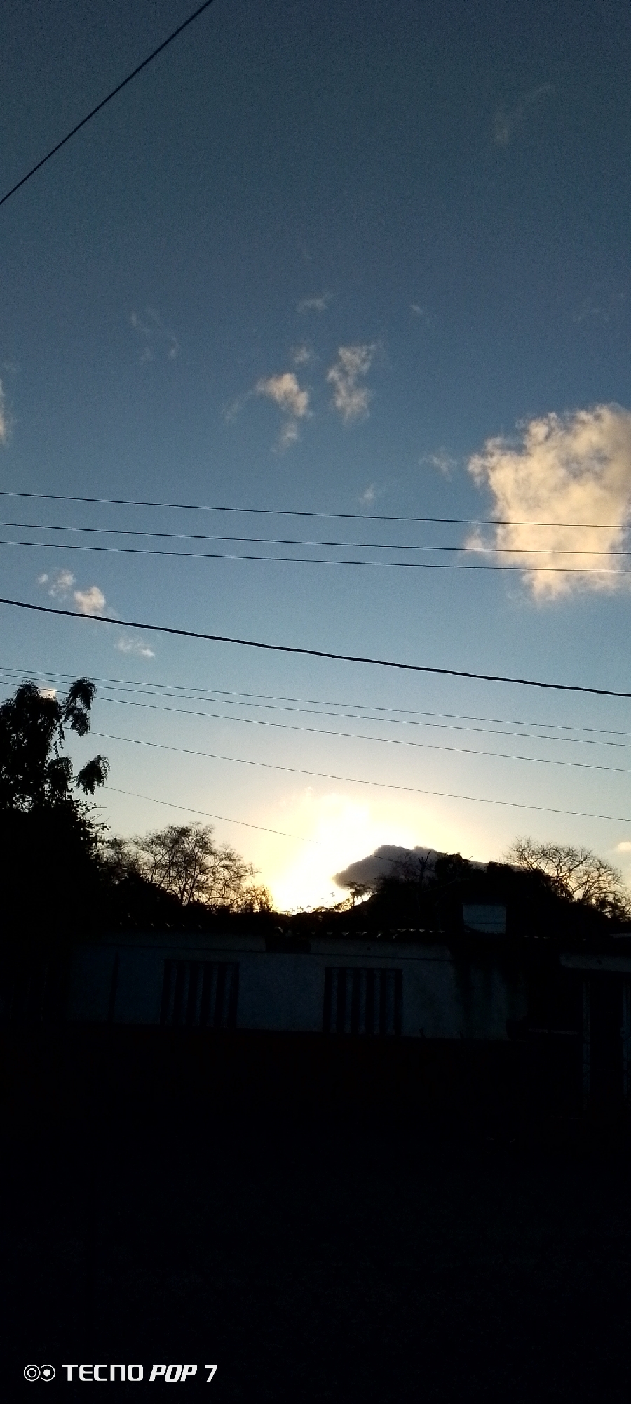 The image depicts a sunset in Altagracia, Venezuela. Silhouetted against the bright sun are trees, a building with barred windows, and utility wires stretching across the sky. Scattered clouds are visible in the upper portion of the image. The lower portion of the scene is dark, obscuring details of the foreground. A "TECNO POP 7" watermark is visible in the bottom left corner.