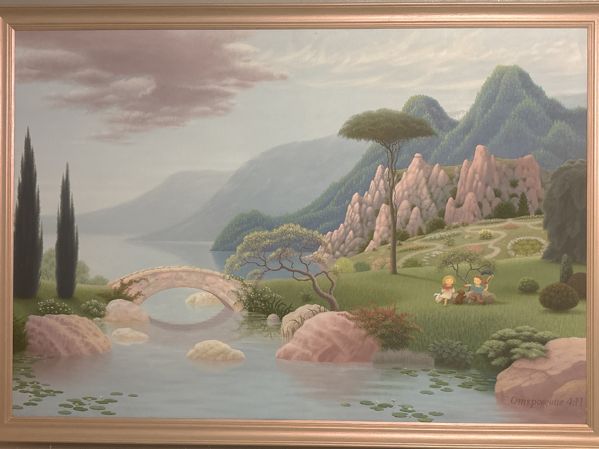 This is a painting depicting a serene landscape. In the foreground, a calm body of water with lily pads is visible, with several large, pinkish rocks scattered near its edge. A stone bridge arches over the water in the middle ground. Beyond the bridge, rolling green hills are dotted with trees and pinkish rock formations, leading up to a series of larger, verdant mountains in the background under a soft, pinkish-grey sky with scattered clouds. In the mid-ground, on a grassy area, two small figures, a boy and a girl, are seated on the grass. The boy is wearing a blue shirt and brown shorts, and the girl is in a white dress. They are interacting with a small brown dog. The scene appears to be set in a park or garden. In the bottom right corner of the painting, there is text that reads "Откровение 4:11".