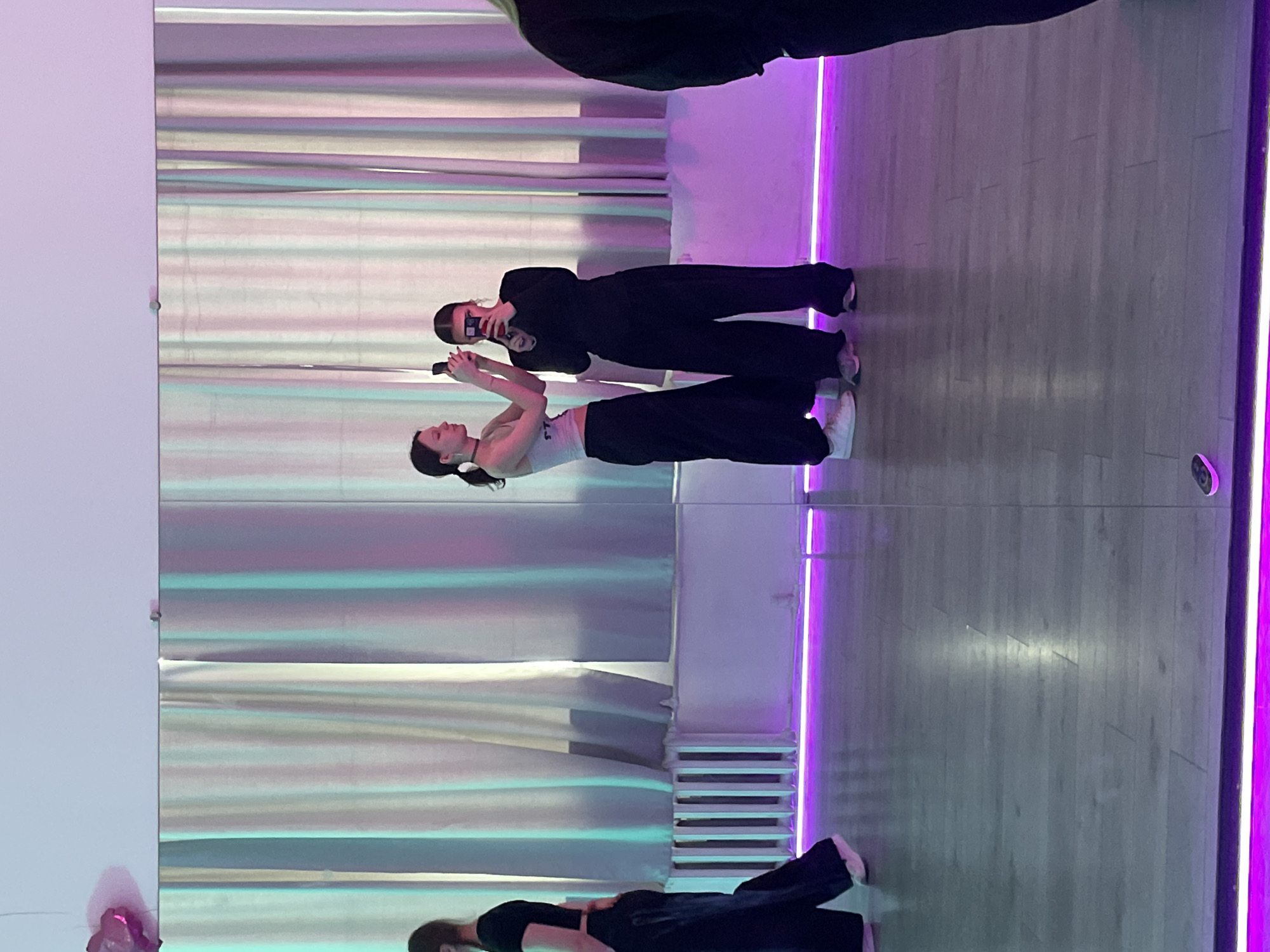 This indoor photograph, taken in Saint Petersburg, Russia, captures a scene primarily through a large mirrored wall. The image features the reflections of two women, seemingly in a studio or dance space.
The woman closer to the foreground wears a light-colored top, dark wide-leg pants, and white sneakers, with her hair tied back. She is holding a smartphone in both hands, angled as if taking a picture or video of herself in the mirror. Behind her, another woman is reflected, dressed in a black long-sleeved top and dark wide-leg pants. She also holds a smartphone, appearing to take a selfie or record the scene. Both individuals are focused on their devices.
The room's interior is visible through the reflection: a light-colored wooden floor and floor-to-ceiling curtains with a horizontal striped pattern that appear iridescent due to the lighting. A white radiator or heating unit is visible on the wall behind the curtains. Bright purple LED strip lights run vertically along the edges of the mirrored wall and possibly along the floor, casting a vibrant glow throughout the space and influencing the color of the reflections. A small, dark security camera or lens is visible in the upper right corner of the mirror's reflection.
Part of a third person (likely the actual photographer) is visible in the top portion of the original image, and a partial reflection of a fourth individual (head and shoulder in dark clothing) can be seen in the bottom left of the mirror, adjacent to a small, indistinct pink object. The artificial lighting suggests the activity is taking place indoors, possibly in the evening or at night. No visible text is discernible in the image.