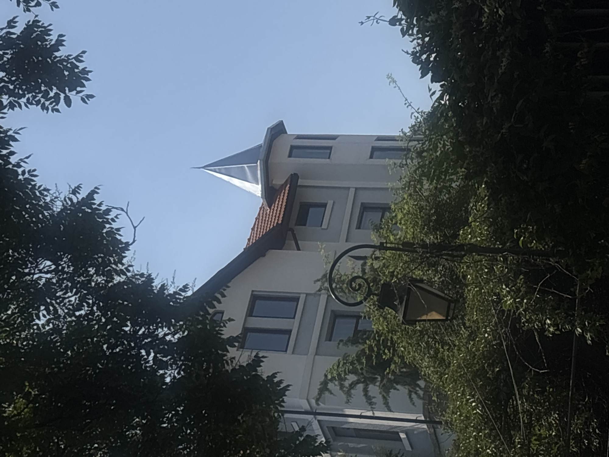 An upward-angled, tilted photograph showcases a distinctive building with a prominent turret against a clear blue sky. The scene is set outdoors, likely in Plovdiv, Bulgaria, under bright daylight conditions.
The building features a sharp, metallic, conical spire or turret at its apex, leading down to light-colored, possibly white or light gray, walls. Multiple rectangular windows are visible across the building's facade. Parts of the roof are covered with reddish-brown tiles, contrasting with the lighter walls.
Lush, dark green foliage, including trees and possibly climbing vines, heavily frames the building on both the left and right sides, partially obscuring the lower architectural details. On the right side, a decorative black wrought-iron street lamp with intricate scrollwork and a glass lantern is visible, partially intertwined with the surrounding greenery.
The time of day appears to be sunny morning or early afternoon, indicated by the bright sky and clear lighting. No people or visible text are present in the image. The perspective suggests the photo was taken from a lower elevation, looking up at the building through the surrounding vegetation.