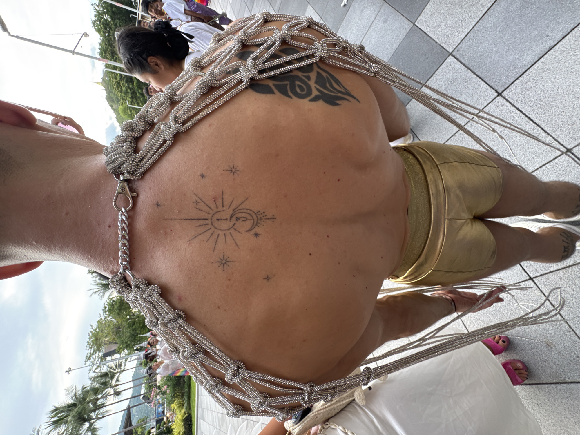 A person's back and shoulders are the primary focus of this image, adorned with a macrame-style harness made of silver beads and chains. The harness is intricately woven and drapes across the person's upper back, secured by a clasp. Two tattoos are visible on the person's back: a large tribal-style tattoo on the left shoulder blade and a celestial design, featuring a sun, moon, and stars, on the mid-back. The person's skin appears tanned.
The person is wearing shiny gold shorts. The background shows a tiled floor, suggesting an outdoor setting. In the blurred background, several people are visible, some wearing white tops, and palm trees indicate a warm climate. The lighting suggests it is daytime, with overcast skies. The overall impression is one of a festive event, possibly a parade or celebration, given the attire and the presence of other people. No text is visible in the image.