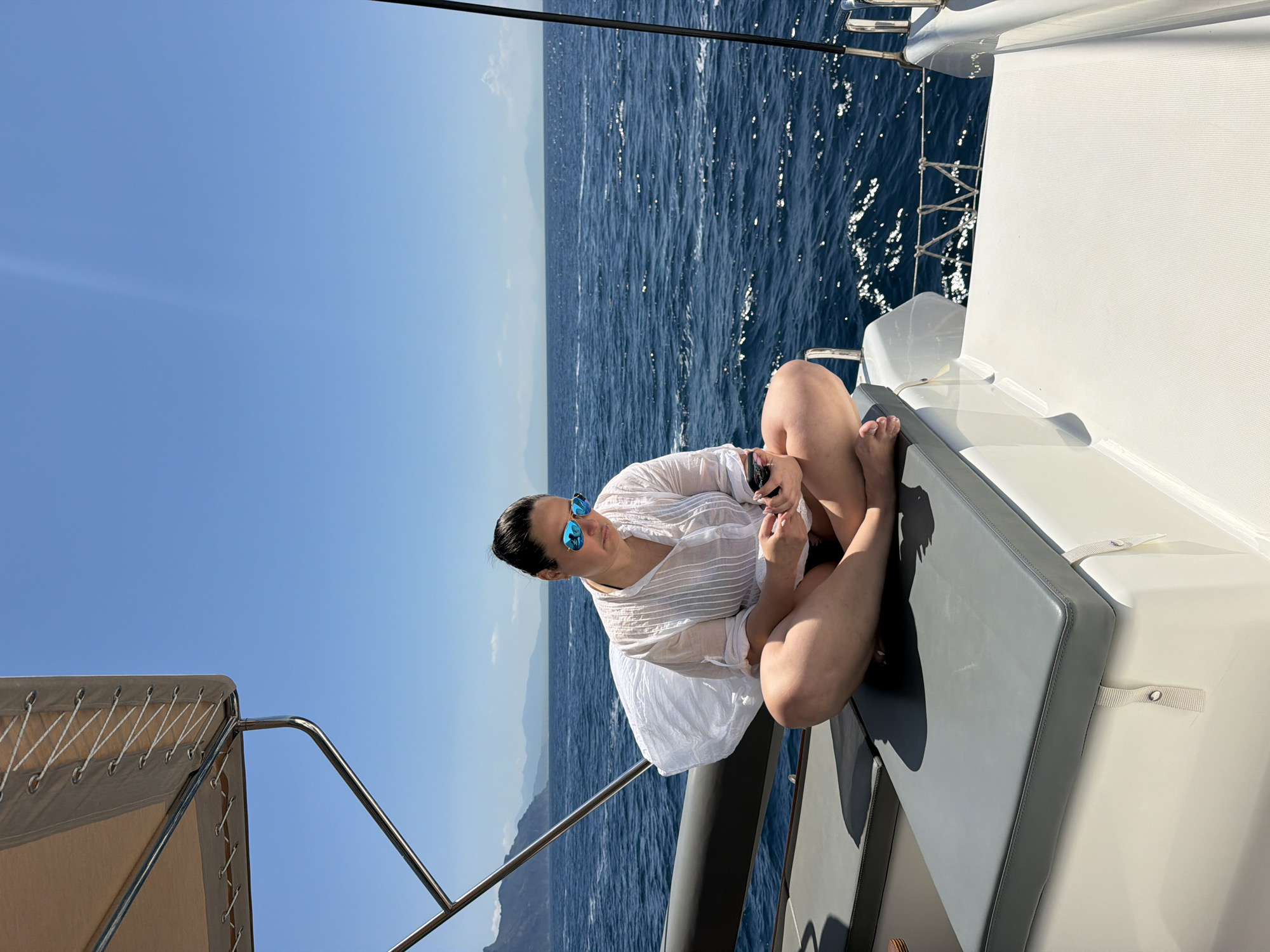 The image shows a woman sitting cross-legged on a boat with blue water and sky in the background. She is wearing a light-colored, sheer, button-down shirt and blue sunglasses. She appears to be holding a device, possibly a phone, in her hands. The boat has a gray cushion where she is sitting and white railings or fixtures. The setting includes a body of water, possibly the sea, and a clear blue sky. There is a distant landmass or island visible on the horizon. The location is not discernible from the image.