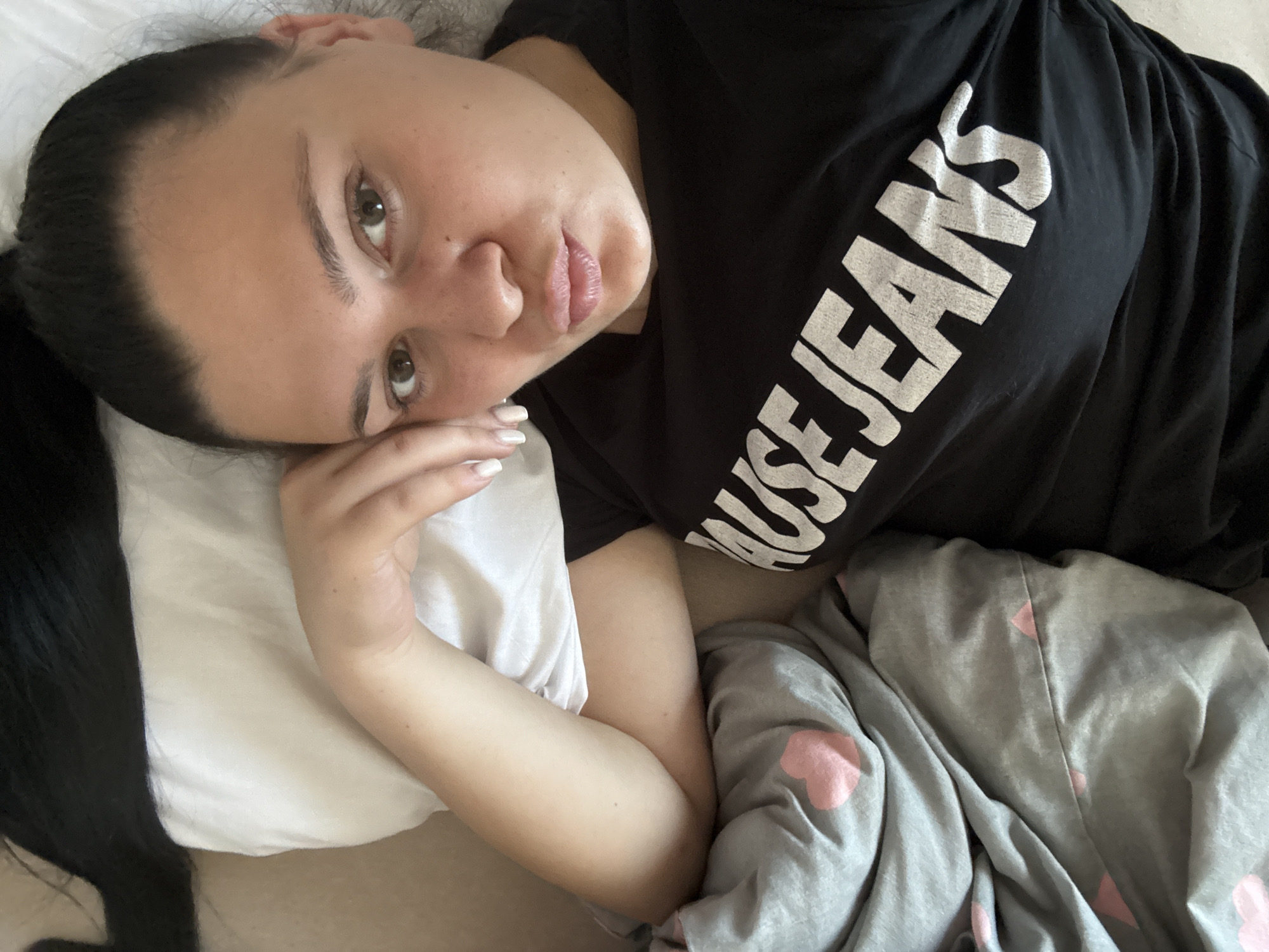 A young woman lies on her side on a bed, her head resting on a white pillow. She has dark hair pulled back and is wearing a black t-shirt with white text that reads "HOUSE JEANS" across the chest. A gray blanket with pink heart patterns is draped over her. The scene is an indoor setting, likely a bedroom, located in Rogosh, Bulgaria.