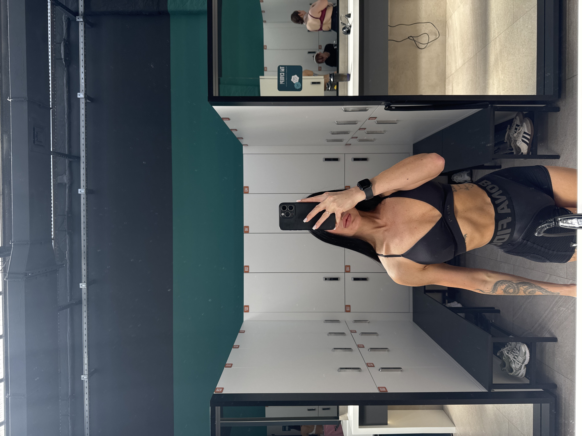 The image depicts a woman taking a mirror selfie in what appears to be a gym locker room or changing area. The woman, shown from the waist up, holds a black smartphone to capture her reflection. She has dark hair, a fit build, and a prominent, detailed tattoo on her left forearm. She is dressed in a black sports bra and black athletic shorts, and wears a black smartwatch on her left wrist.
The setting features rows of white lockers with silver handles and orange numbered labels, such as "123", "124", "141", and "142". A dark grey bench is positioned below the lockers. In the reflection, a pair of athletic shoes, possibly adidas, rests on a lower shelf. The walls are segmented with a dark grey/black area and a distinct dark teal or green section.
Further in the mirror's reflection, another individual wearing a red top is partially visible sitting, suggesting other people are present in the changing facility. A sign is also reflected, displaying reversed text that reads "ЛР КАБИН" (likely "КАБИН ЛР"), which could translate to "Changing Room," along with a blue floral logo. The waistband of the woman's shorts clearly shows the brand "ADIDAS" in reversed text. The floor is tiled with a light, neutral color. The scene is indoors, presumably in a fitness center in Kazan, Russia.