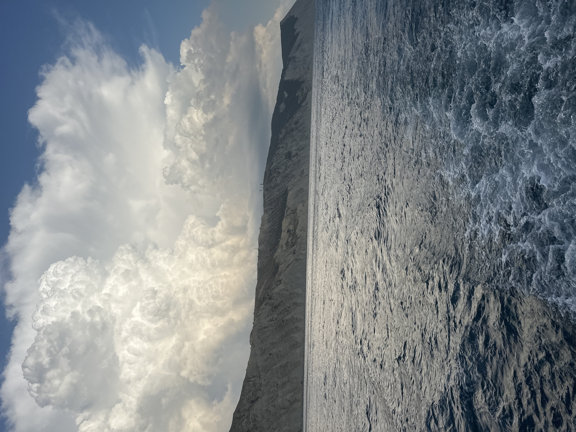 The image shows a view of the Black Sea coast near Anapa, Russia. In the foreground, the dark blue water is choppy, with white foam trailing behind a moving vessel, suggesting the viewpoint is from a boat. The middle ground features a steep, rocky coastline with sparse vegetation. In the background, large, dramatic cumulonimbus clouds dominate the sky, with a sliver of blue sky visible to the right. The lighting suggests it is daytime, possibly late afternoon or early morning, with sunlight illuminating the clouds. There are no people or specific structures immediately visible on the coast, though a faint path or road appears to run along the cliff top, and there are a few indistinct vertical structures on the ridge.