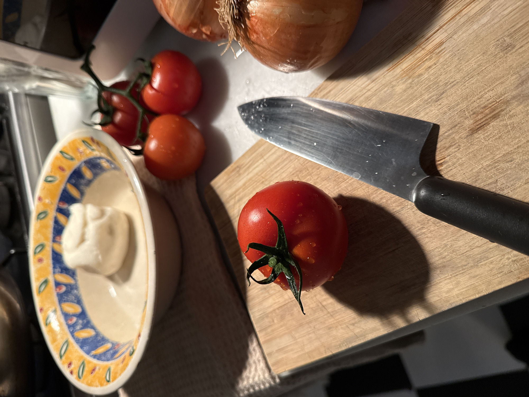 The scene is a still life of food preparation. A wooden cutting board sits on a surface with a black and white checkered pattern. On the cutting board, there is a red tomato with a green stem and a chef's knife with a black handle. Next to the cutting board are two brown onions, three more tomatoes on a vine, and a bowl with an ornate blue and yellow pattern containing a white solid. The lighting is bright, and the surfaces appear wet, possibly from washing. The image does not show people or details that would indicate it is located in Barcelona, Spain.