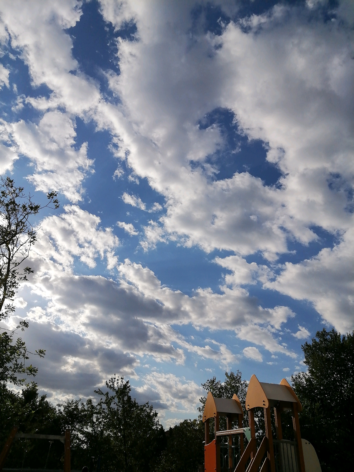 The image captures a playground scene with a dramatic sky overhead. The sky is a vibrant blue, filled with a mixture of white and grey clouds that appear to be moving. Sunlight filters through some of the clouds, creating highlights and shadows. In the foreground, the top sections of a children's playground structure are visible. These structures are made of colorful materials, including orange and beige panels, with wooden beams and grey metal accents. To the left and right of the playground equipment, dense green trees form a natural backdrop. The overall impression is of a bright, possibly late afternoon or early evening, day in a park or recreational area. The specific location of Rostov-on-Don, Russia, is not visually discernible from the image itself.