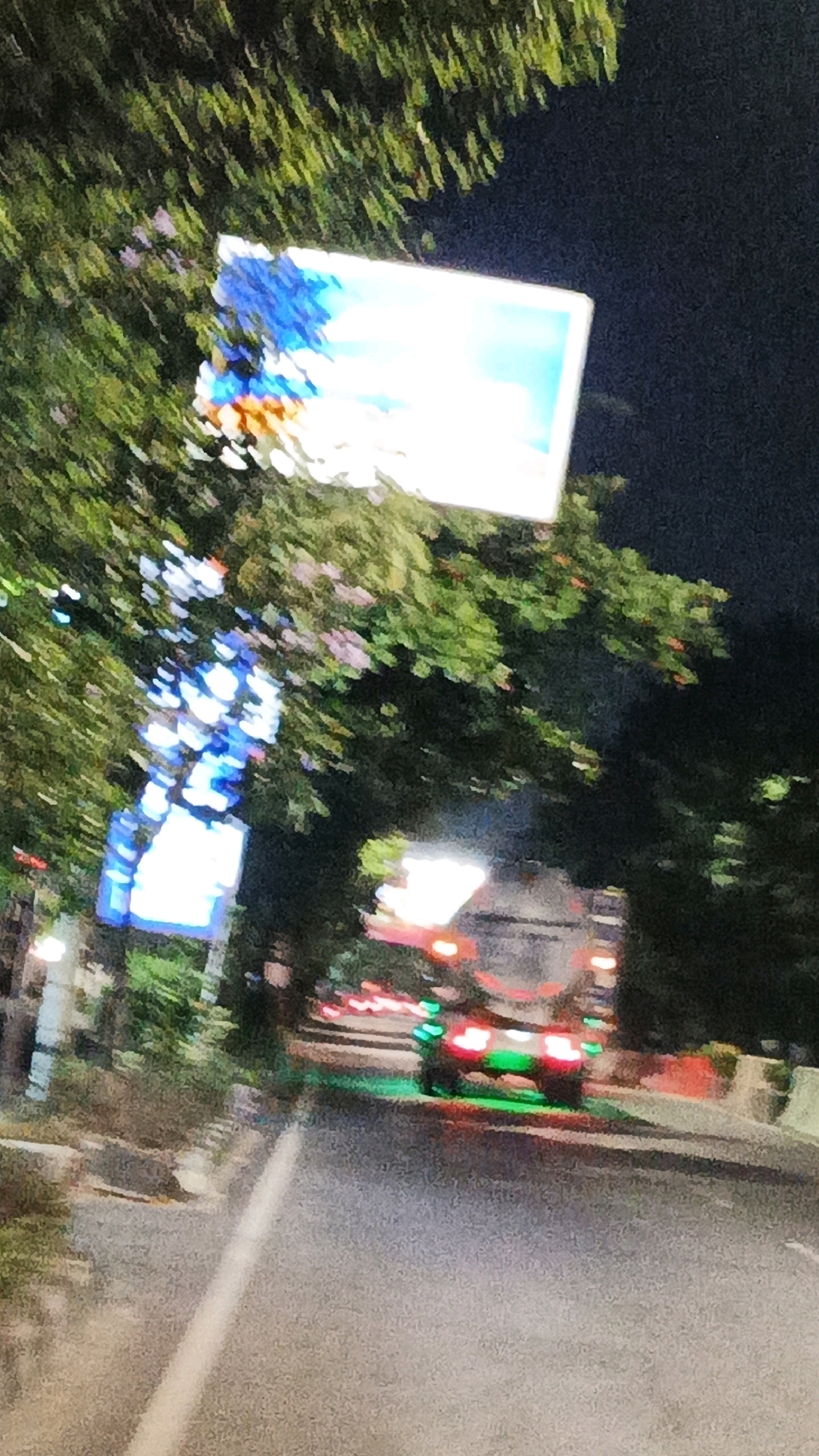 This is a nighttime, out-of-focus, vertical-format photograph taken from a moving vehicle on a street in Surakarta, Indonesia. A large, rectangular billboard with a blurry image of blue and white shapes is visible in the upper left quadrant, partially obscured by tree branches. To the left of the billboard and slightly lower, a vertical structure illuminated with blue lights, possibly a decorative pole or another sign, is also visible. In the middle ground, a large truck with its brake lights illuminated in red and green is driving away from the camera. The road surface is visible in the foreground, with a white lane marker. The overall environment appears to be a city street at night, with streetlights and the illuminated elements creating a somewhat vibrant but blurred scene.