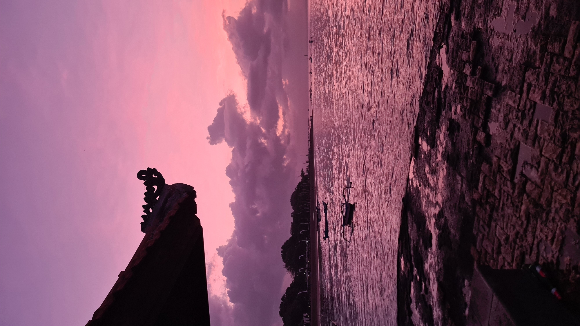 This is a vertical, eye-level shot of a beach in Denpasar, Indonesia at dusk. The sky is a vibrant gradient of pink and purple, with dramatic, dark clouds. The water is a deep pink, reflecting the sky. In the foreground, there are dark, wet rocks. A traditional Balinese roof overhang with intricate carvings is visible in the upper left corner. Several traditional Balinese fishing boats are scattered on the water. In the middle ground, there is a tree-lined shore with some structures that appear to be part of a resort or buildings. The scene is serene and picturesque, characteristic of a tropical sunset.
