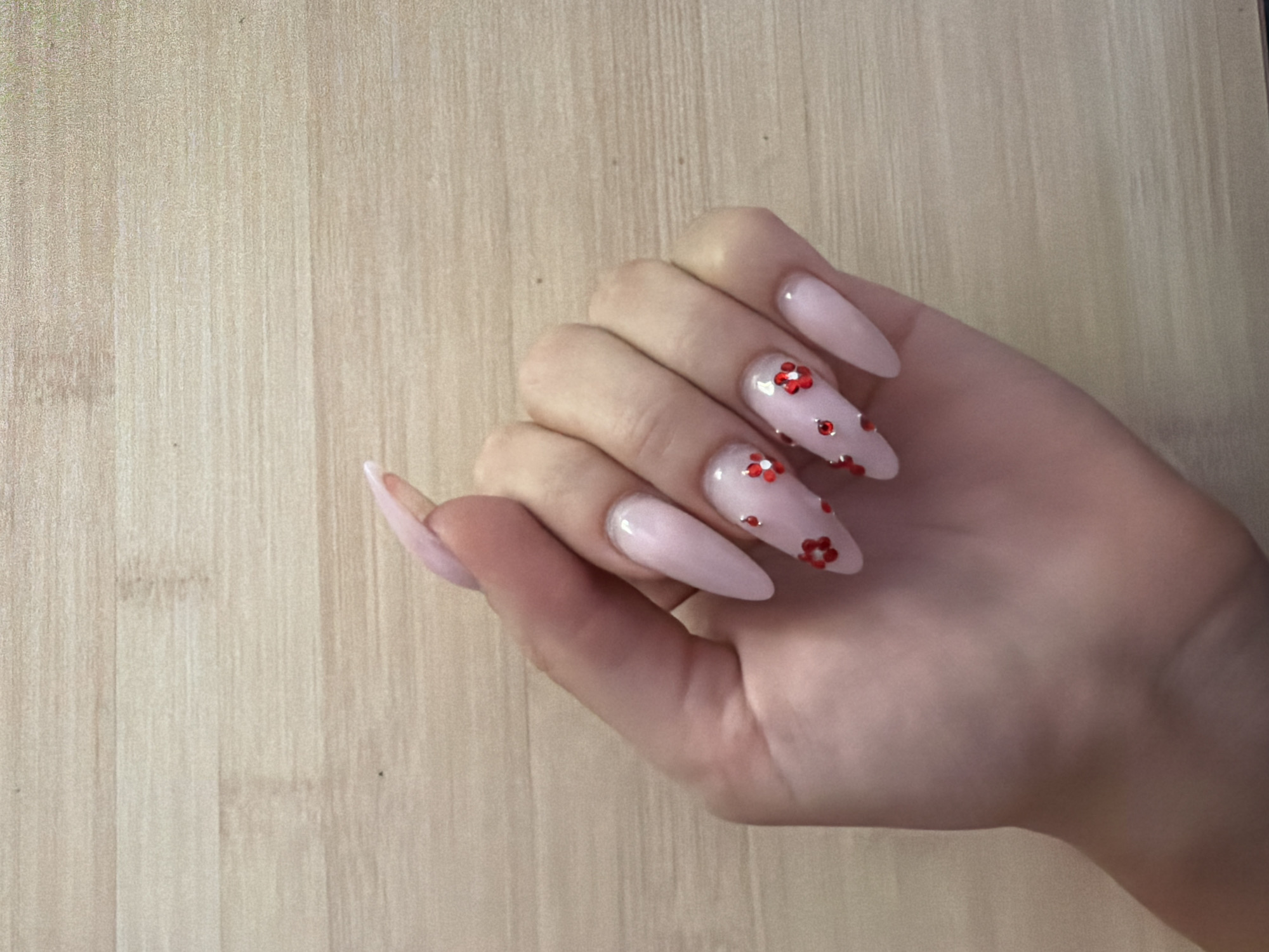 The image shows a person's hand with stiletto-shaped fingernails painted in a pale pink color. The nails are decorated with small red flower-shaped embellishments. The hand is resting on a light brown wooden surface, possibly a table or desk. There is no indication of the city of Prosena, Bulgaria, or any other specific location within the image. The lighting suggests an indoor setting.