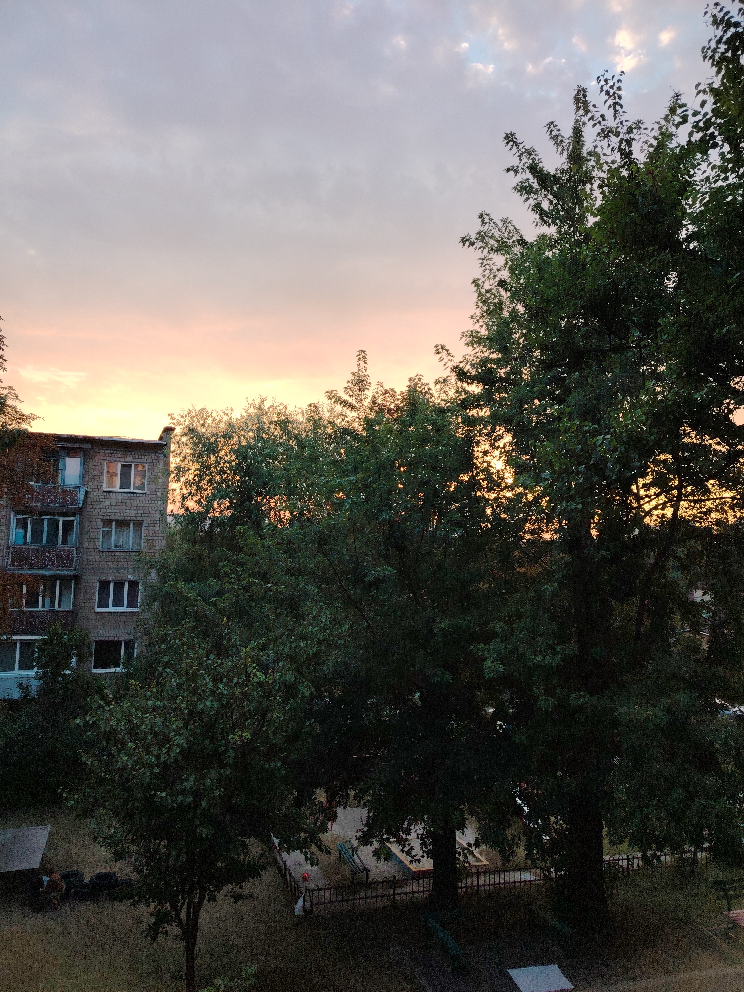The image depicts an apartment building with multiple stories and balconies, situated amidst a cluster of trees, with a sky displaying pastel hues of pink and orange, indicative of a sunset or sunrise. The building's facade is constructed of brick, and the windows are of varying sizes and designs, some with closed shutters. Lush green foliage from the trees partially obscures the building and the sky, suggesting an urban or suburban setting. In the foreground, a playground area with benches and a fence is visible, along with some tires and a small shelter. The overall scene conveys a sense of quietude and the transition between day and night. The location is identified as Kyiv, Ukraine.