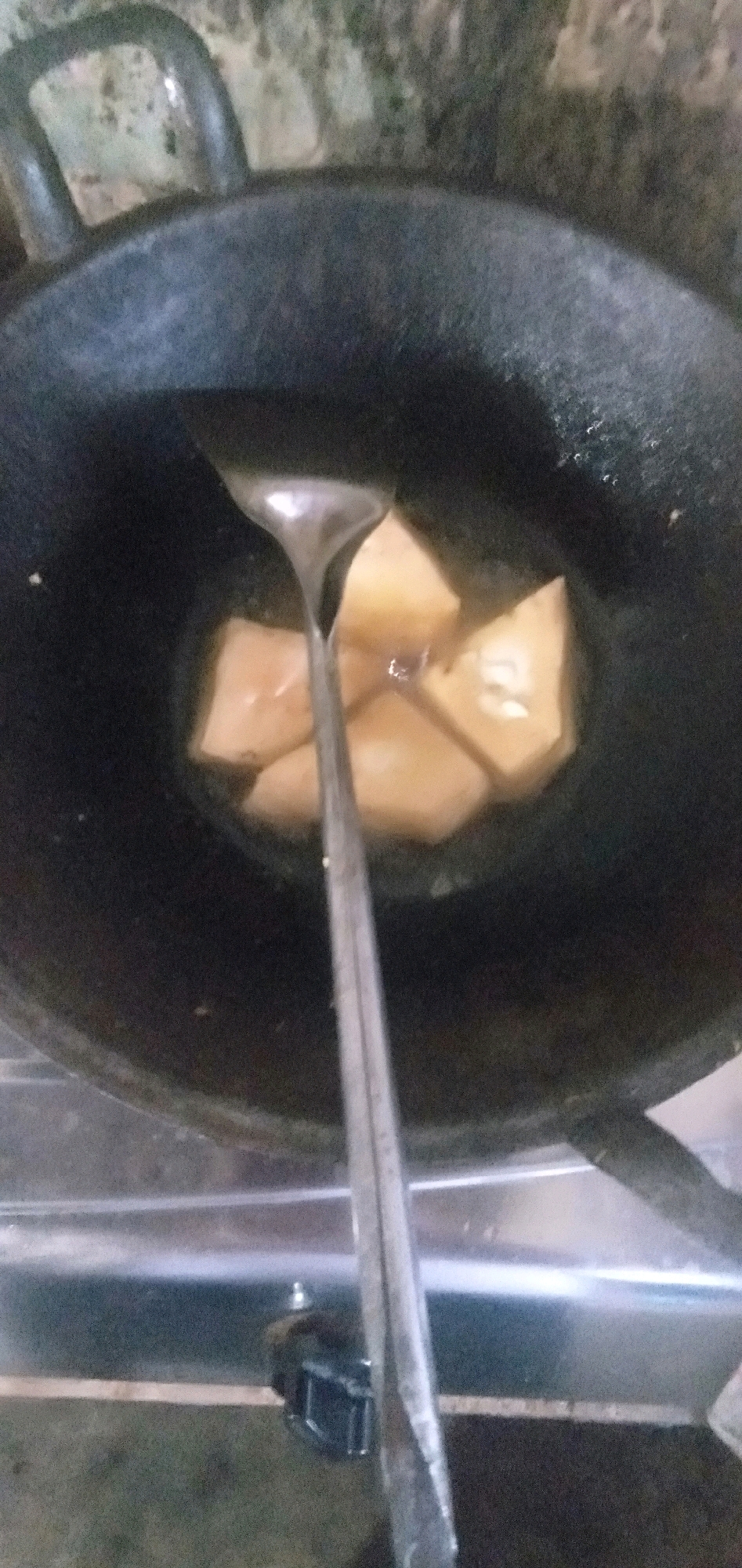 The image shows a close-up, top-down view of a black wok with several pieces of light brown food, likely fried tofu or tempeh, submerged in oil. A metal spatula is positioned over the food, with its head partially submerged in the oil. The wok is on a stovetop. The background is out of focus, showing a textured, light-colored surface and parts of the stovetop. The overall scene suggests food preparation or cooking. The image does not contain enough information to determine its specific location within Plemahan, Indonesia.