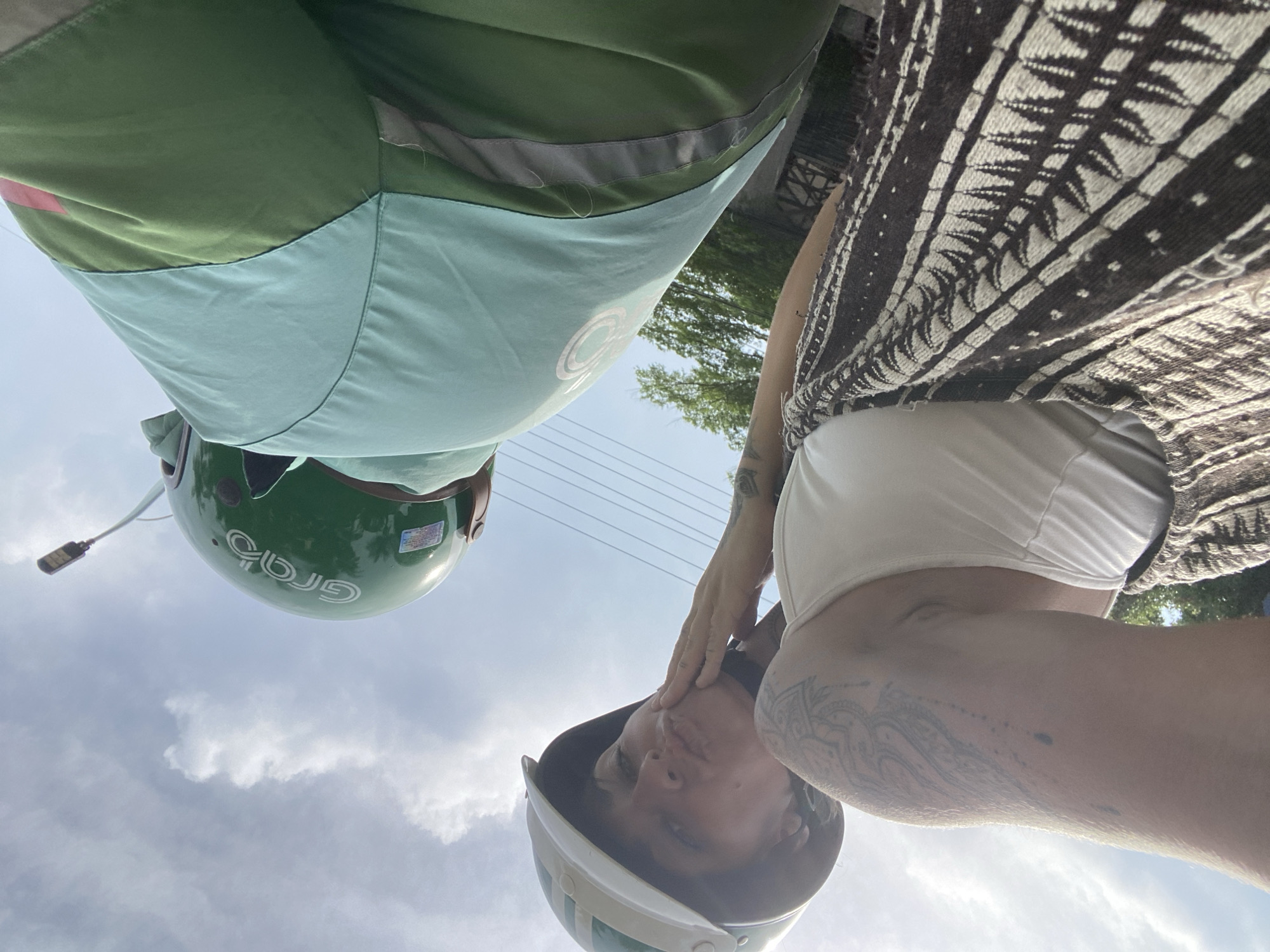 The image shows two people on a Grab motorcycle in Phu Thuy Ward, Vietnam. The person in the foreground is a woman wearing a white tank top and a patterned skirt. She is wearing a white and green helmet and has her hand on her chin, making a kissing face towards the camera. She has tattoos on her left arm. The second person, presumably the Grab driver, is wearing a green helmet with the Grab logo and a light blue and green uniform. The background shows a blue sky with clouds and some greenery. The image is taken from a low angle, looking up at the sky.