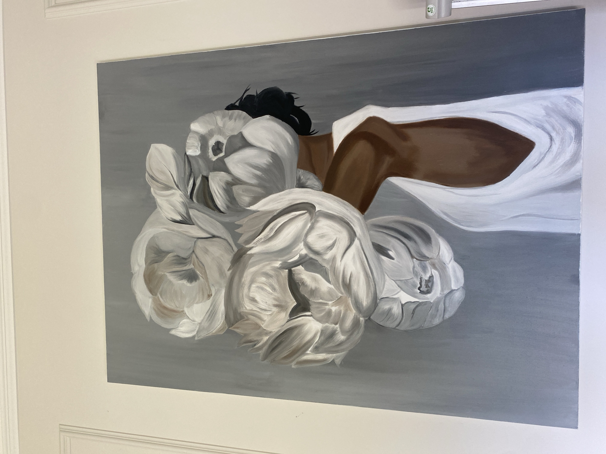 A large, vertical painting is displayed on a light-colored wall. The painting features a woman with dark skin, wearing a white dress. Her face is obscured by a large arrangement of white peonies, depicted in varying stages of bloom. The background of the painting is a solid, muted gray. The wall on which the painting hangs is white and features white decorative molding along the top edge. To the right of the painting, a small, white label with green writing is affixed to the wall. This painting is located in the city of Buk, Poland.