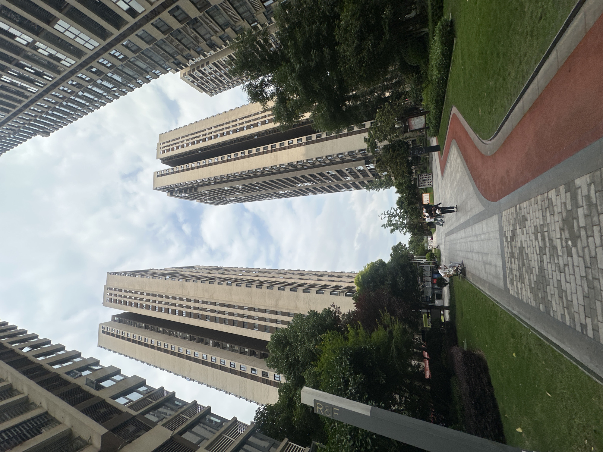 The image depicts a street scene in the Chongqing High-tech Zone, China. Tall apartment buildings dominate the skyline, angled upwards from the perspective of the photographer. A paved pathway with a red running track border winds through a green lawn. Trees are interspersed between the buildings and along the pathway. People are visible in the distance, walking along the path. A sign marked "R&F" is visible in the foreground. The sky is overcast with some patches of blue.