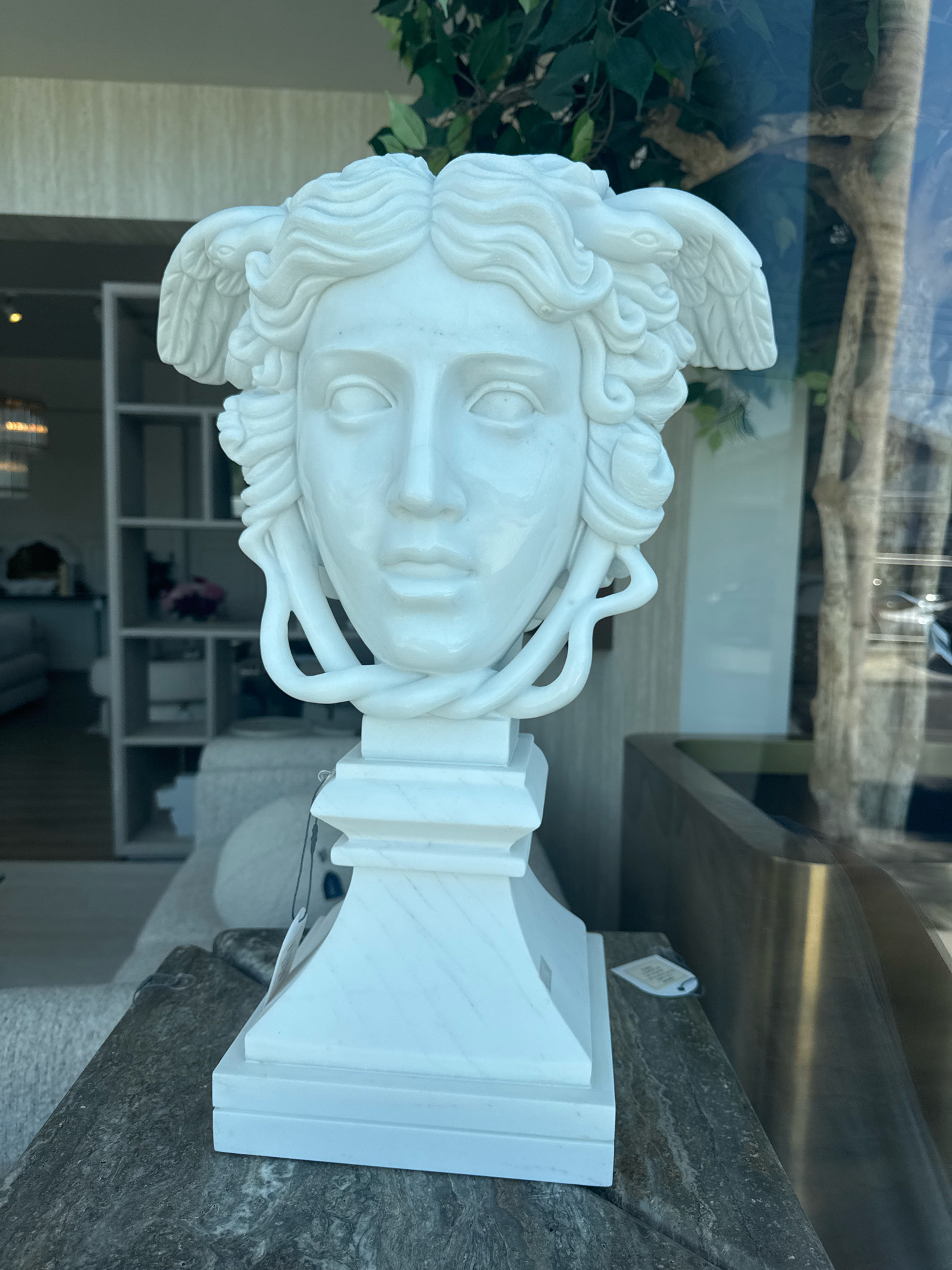 A white marble bust of Medusa, sculpted in a classical style, stands on a marble pedestal. The bust depicts Medusa with serpents intertwined in her hair and small wings on either side of her head, resembling those of a bird. The background shows an indoor setting with furniture and a large window reflecting an outdoor scene. The item appears to be displayed in a showroom or shop in Agios Athanasios Municipality, Cyprus.