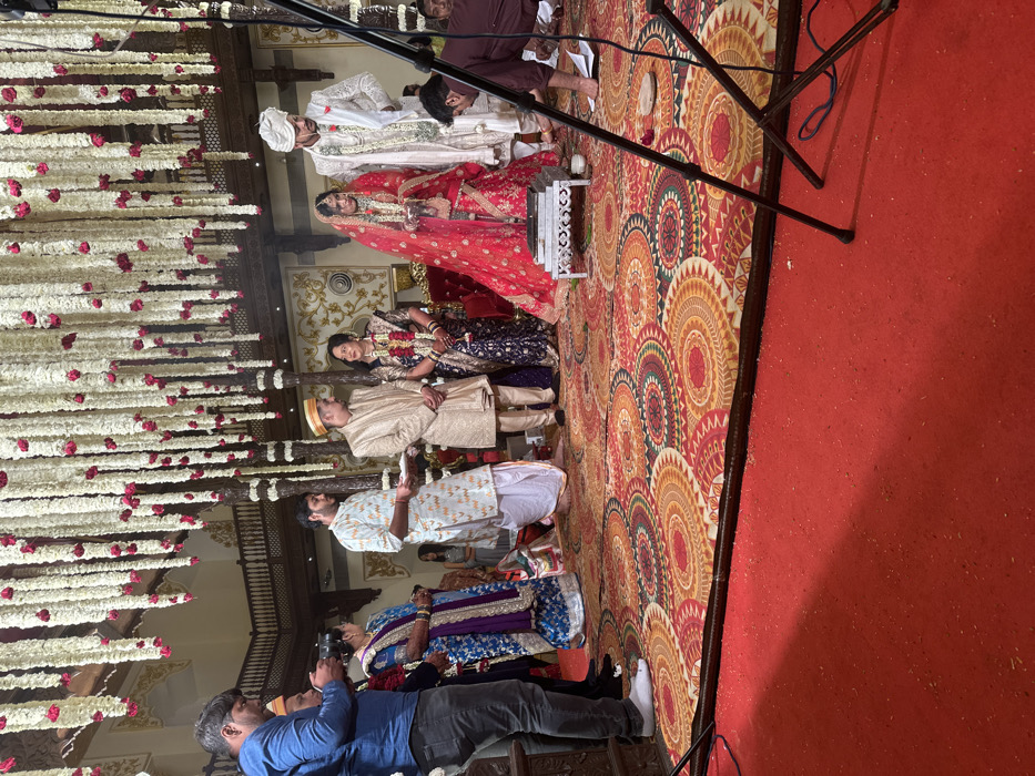 An indoor Indian wedding ceremony is in progress within a grand hall, consistent with the architectural style found at the Lakshmi Vilas Palace in Vadodara, Gujarat, India. The ceiling is densely adorned with hanging garlands of white and red flowers.
At the center of the scene, a bride in a red, heavily embroidered lehenga and veil stands on the right. To her immediate left is the groom, dressed in a white sherwani, white turban, and floral garlands. Between them, a small square fire pit (hawan kund) rests on a white, carved wooden stand, with faint smoke visible.
To the groom's left, an older man in a gold sherwani and yellow turban stands next to an older woman in a dark blue and gold saree, both adorned with garlands. Adjacent to them, a younger man in a white printed kurta and dhoti holds a white tray. On the far right, a man in a maroon shirt crouches on the floor, tending to ceremonial items. On the far left, a man in a blue shirt and grey trousers operates a camera.
The floor of the ceremonial area is covered by a large rug featuring concentric circular patterns in red, orange, gold, and green. The immediate foreground is covered by a plain red carpet. A black tripod stand is partially visible on the lower right. The background features ornate wooden balustrades, pillars, and gilded walls, indicating a richly decorated venue.
