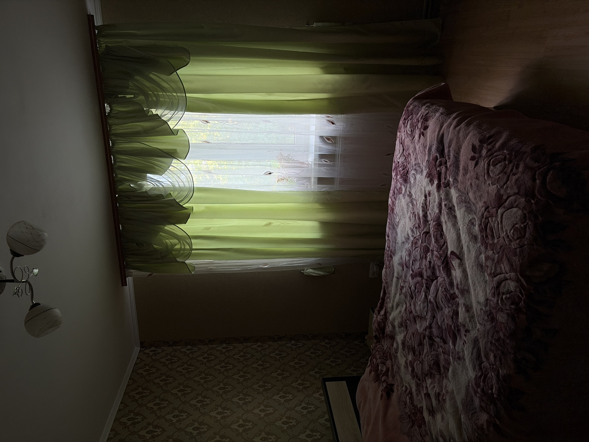 The image shows a dimly lit bedroom. A bed covered with a patterned blanket is the most prominent object in the foreground. The bed has a headboard. To the right, a window is covered by green and white curtains. A ceiling light fixture is visible in the upper left corner. The walls are wallpapered on the left and painted on the right.
