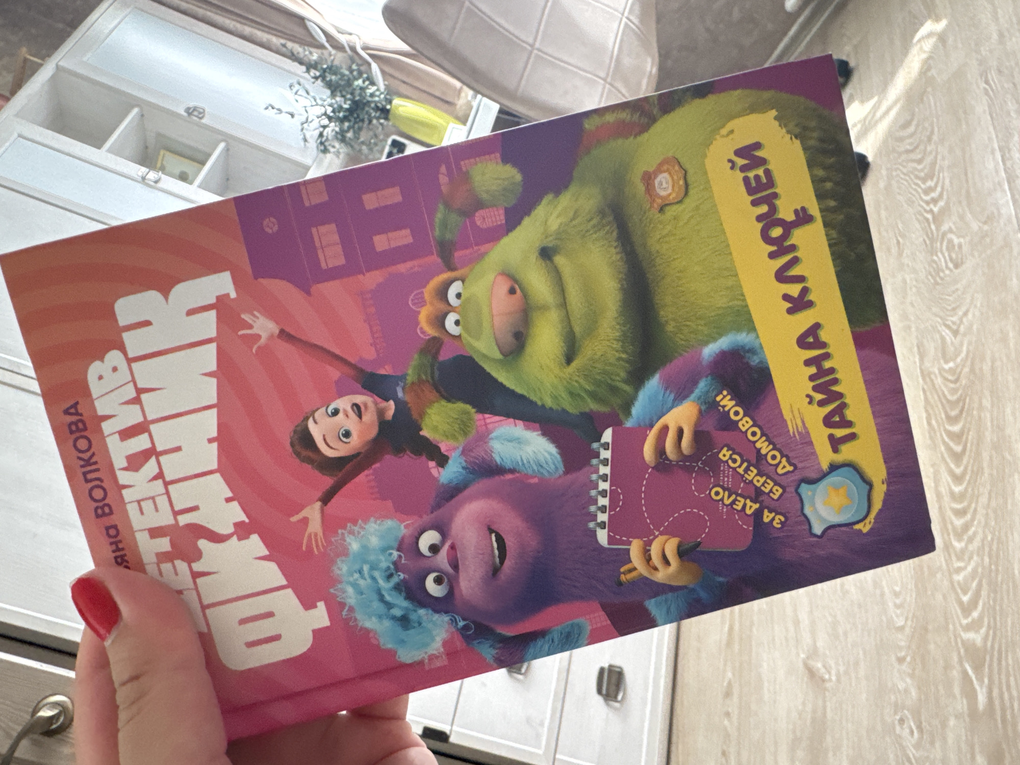 A hand holds a children's book titled "ФИКСИ-ДЕТЕКТИВ" by Яна Волкова. The cover features cartoon-like figures: a green monster, a purple monster holding a notebook, and a girl with brown hair. The title "ТАЙНА КЛЮЧЕЙ" is printed vertically in yellow. The book is held indoors, with furniture visible in the background. There is no indication the location is in Раздолье, Russia.