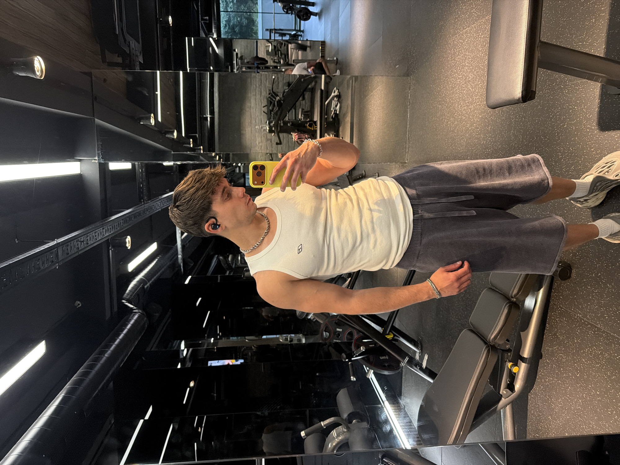 A young man is taking a selfie in a gym. He is wearing a white tank top, gray shorts, white socks, and light-colored shoes. He has a silver chain around his neck and a bracelet on his wrist. He is holding a yellow cell phone. He has a wireless earbud in his left ear. He is standing in front of a mirrored wall with multiple levels. Behind him is a weight bench and other gym equipment. The gym appears to be located in Sofia, Bulgaria, based on the architecture and the style of the gym equipment.